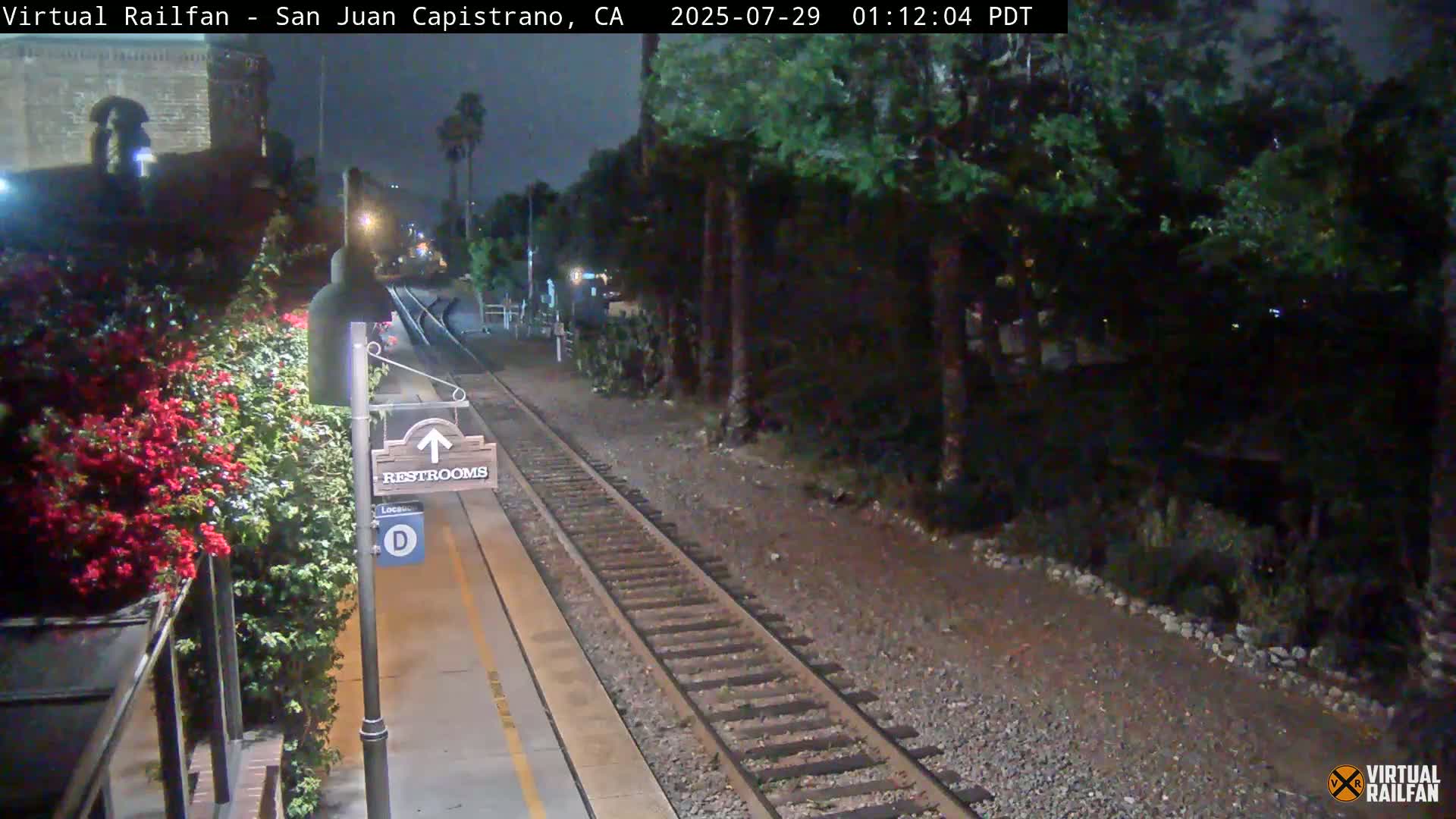 A nighttime view of a train station platform with a restroom sign, railroad tracks, and lush greenery alongside the platform under a dark sky.