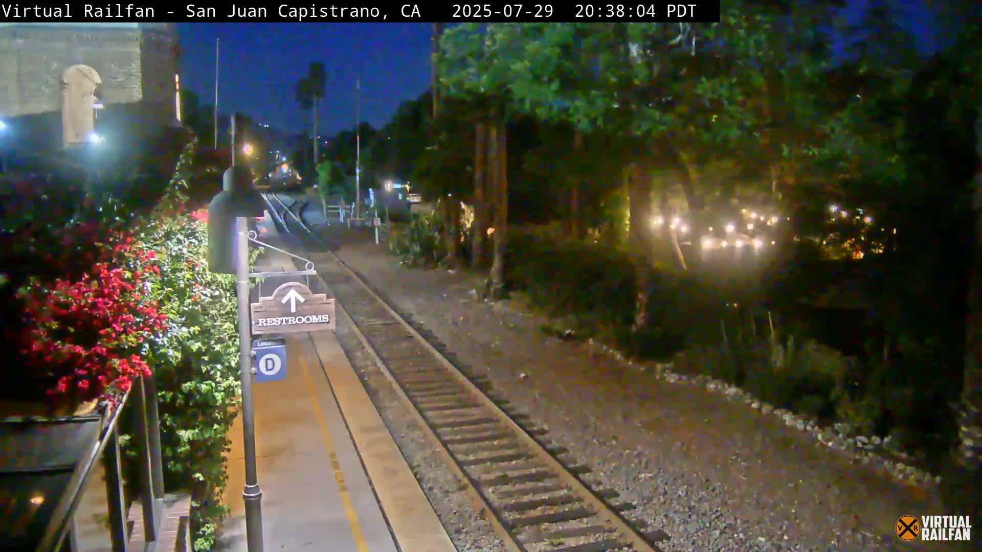 A nighttime view of a train station platform with a restroom sign, railroad tracks, and lush greenery alongside the platform under a dark sky.