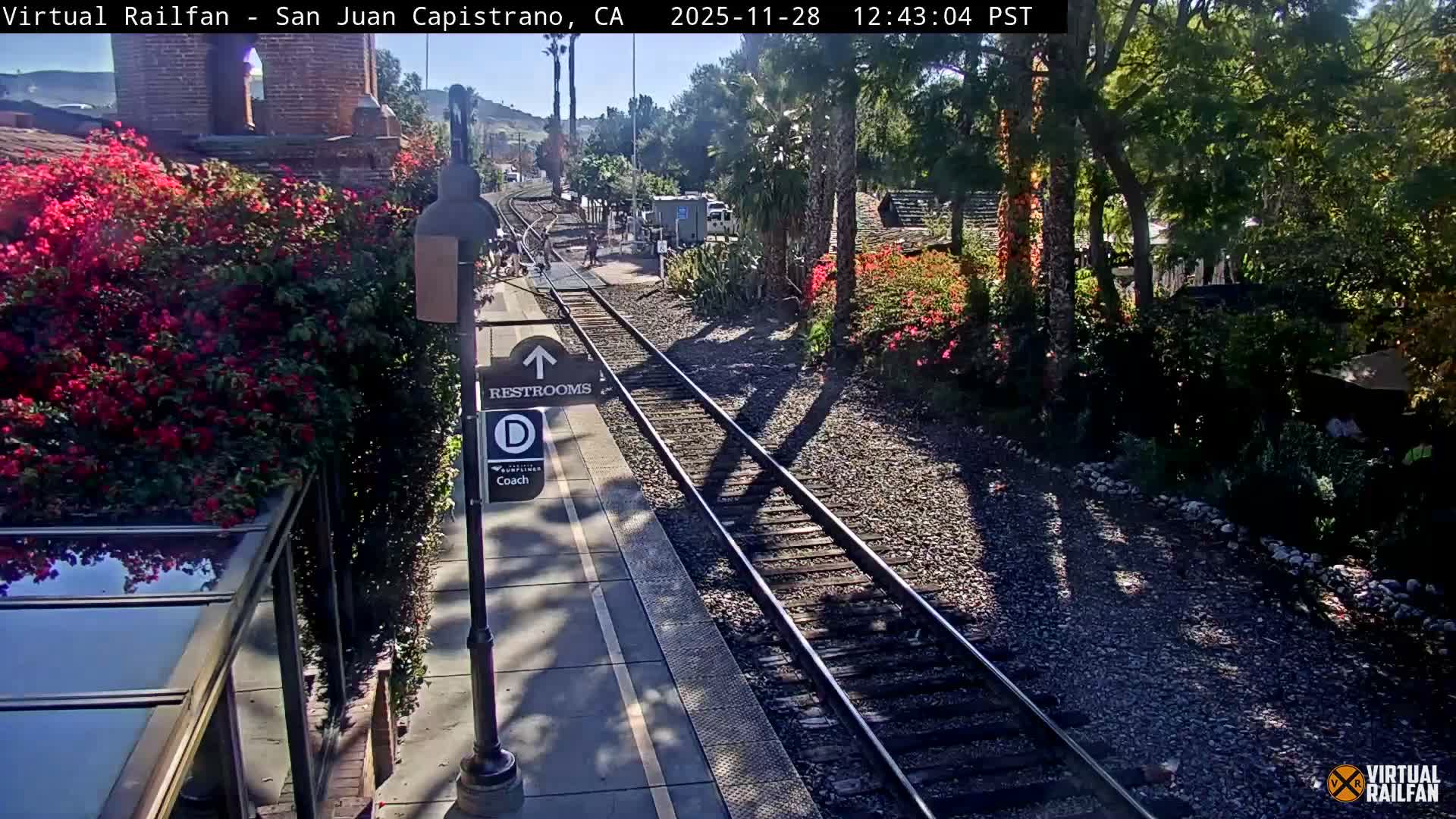 Under a bright, sunny sky, a railway track runs alongside a platform adorned with vibrant red flowering bushes and diverse greenery, with distant people and structures visible.