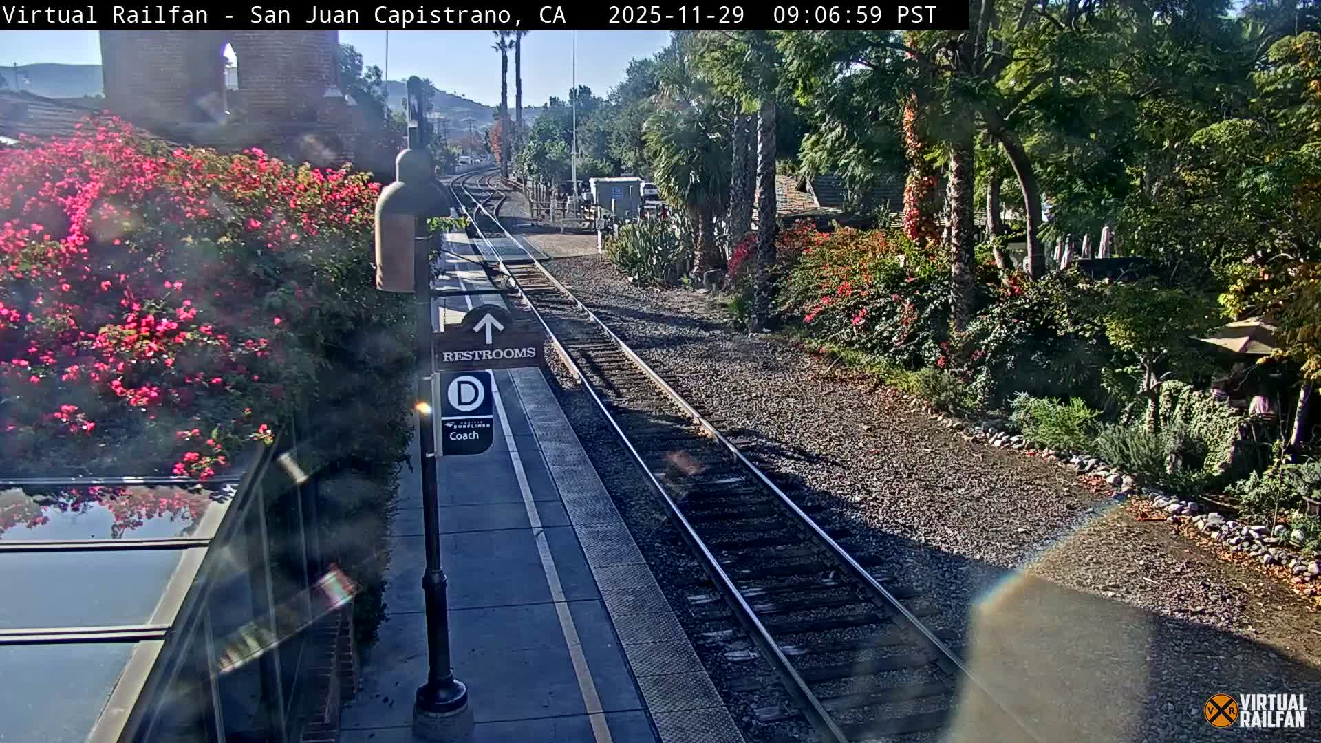 A sun-drenched train station platform and two sets of tracks are bordered by vibrant pink flowering bushes and lush green trees, including palms, under a clear blue sky.