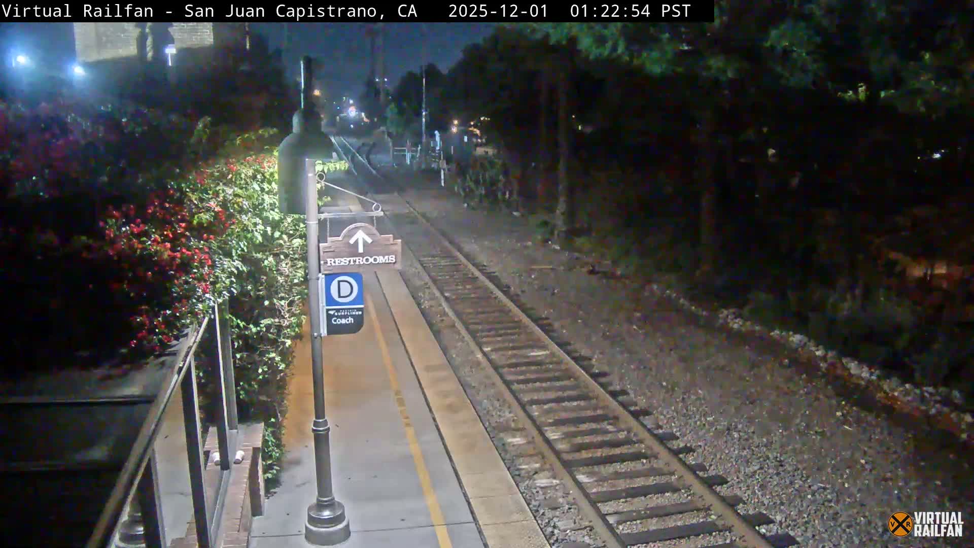 A dimly lit train station platform and railway tracks are visible at night, with a lamppost holding directional signs on the left, lush green and red flowering bushes behind it, and dense trees lining the tracks on the right, all under clear night conditions.