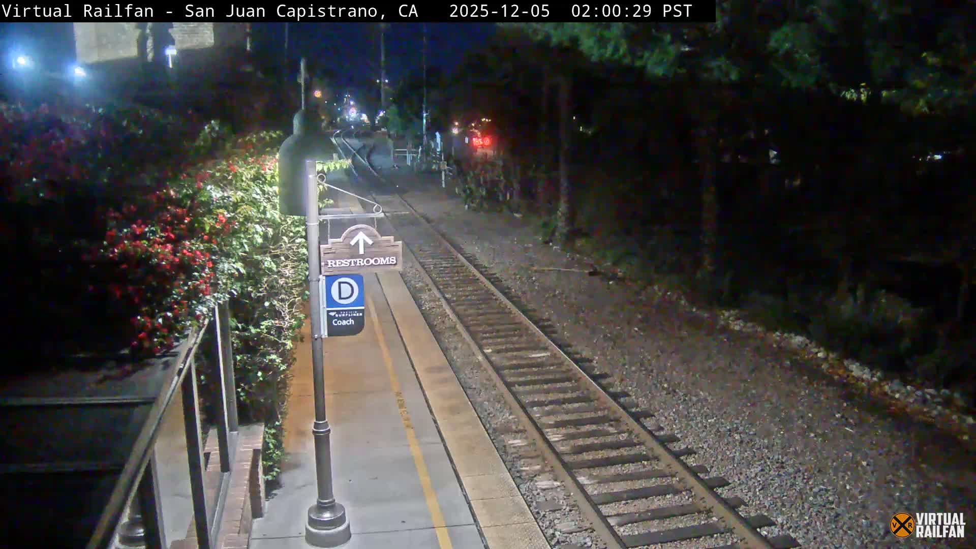 A nocturnal view shows a brightly lit train platform alongside railway tracks, featuring a sign for restrooms and coach "D," flanked by lush greenery with some red flowers, under clear weather conditions.
