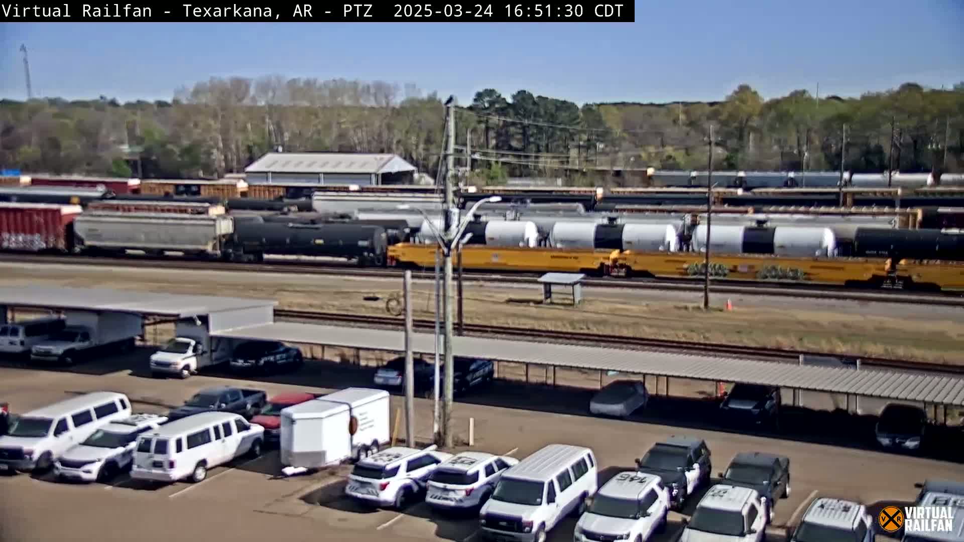 A long freight train passes by a parking lot filled with mostly white SUVs and limousines under a covered structure, on a sunny day.