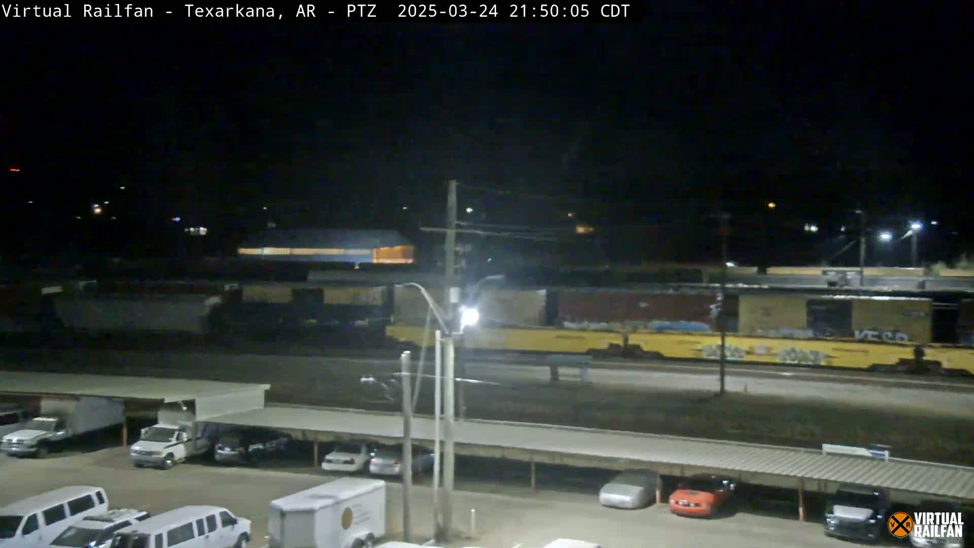 A nighttime view of a freight train passing by a parking lot with several cars and vans under a covered structure.