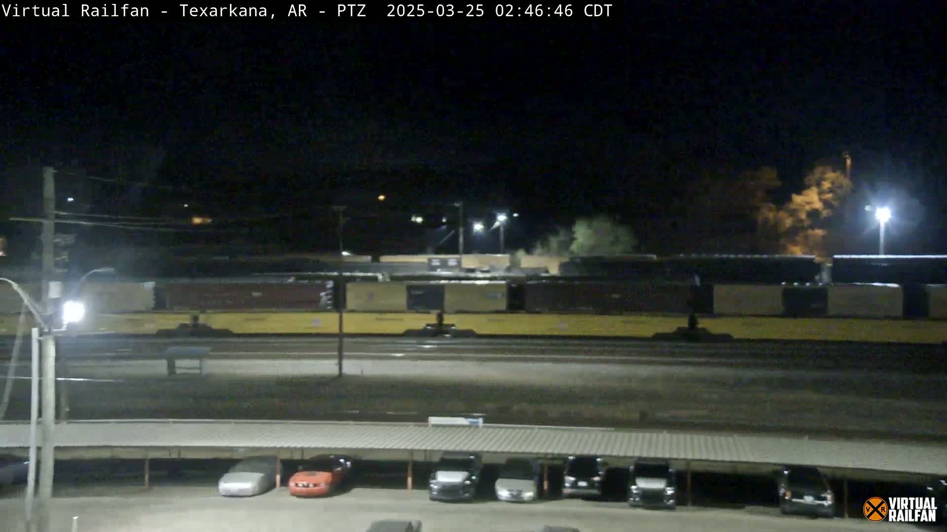 A long freight train sits stationary at night under a dark sky next to a parking lot with several cars.