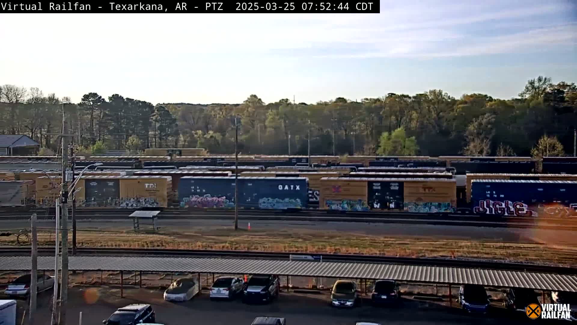 A long line of graffiti-covered train cars sits in a yard next to a parking lot under a covered structure, on a partly cloudy day.