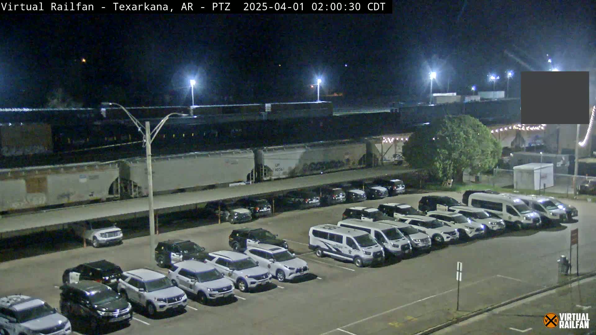 A nighttime view of a parking lot filled with mostly white vehicles, next to a train parked under a covered structure.