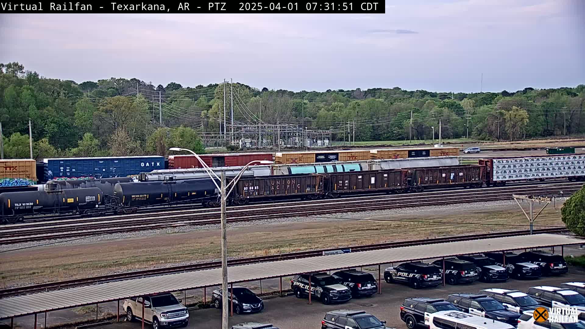A long freight train sits on railroad tracks under a partly cloudy sky, with a parking lot full of police cars visible in the foreground.