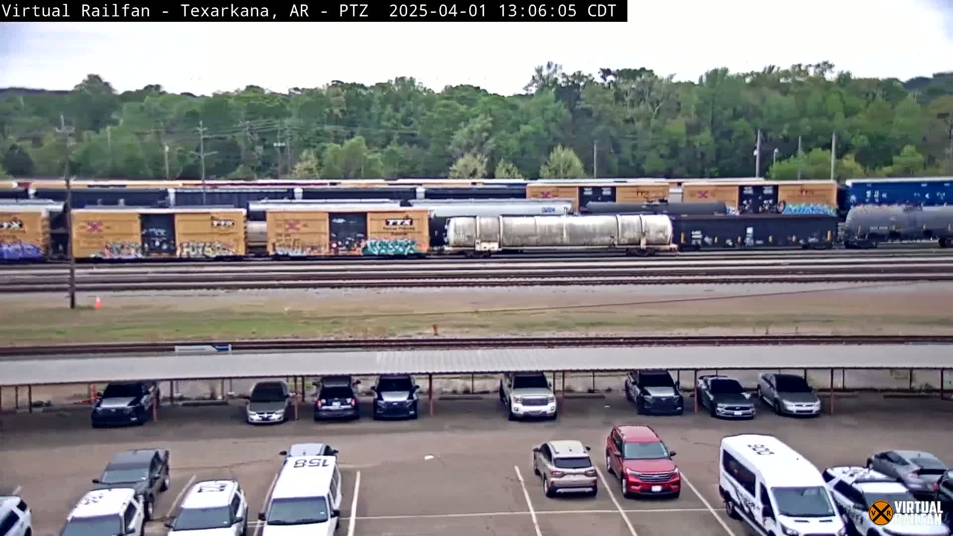 A long freight train sits on railway tracks behind a parking lot with cars parked under a covered structure, all under a partly cloudy sky.