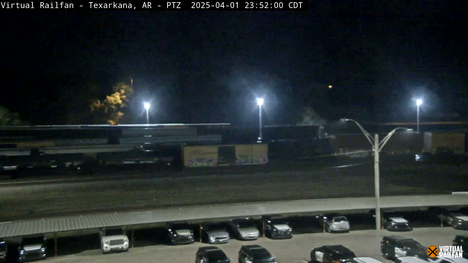 A nighttime view shows a long freight train parked under bright lights, next to a parking lot full of cars.