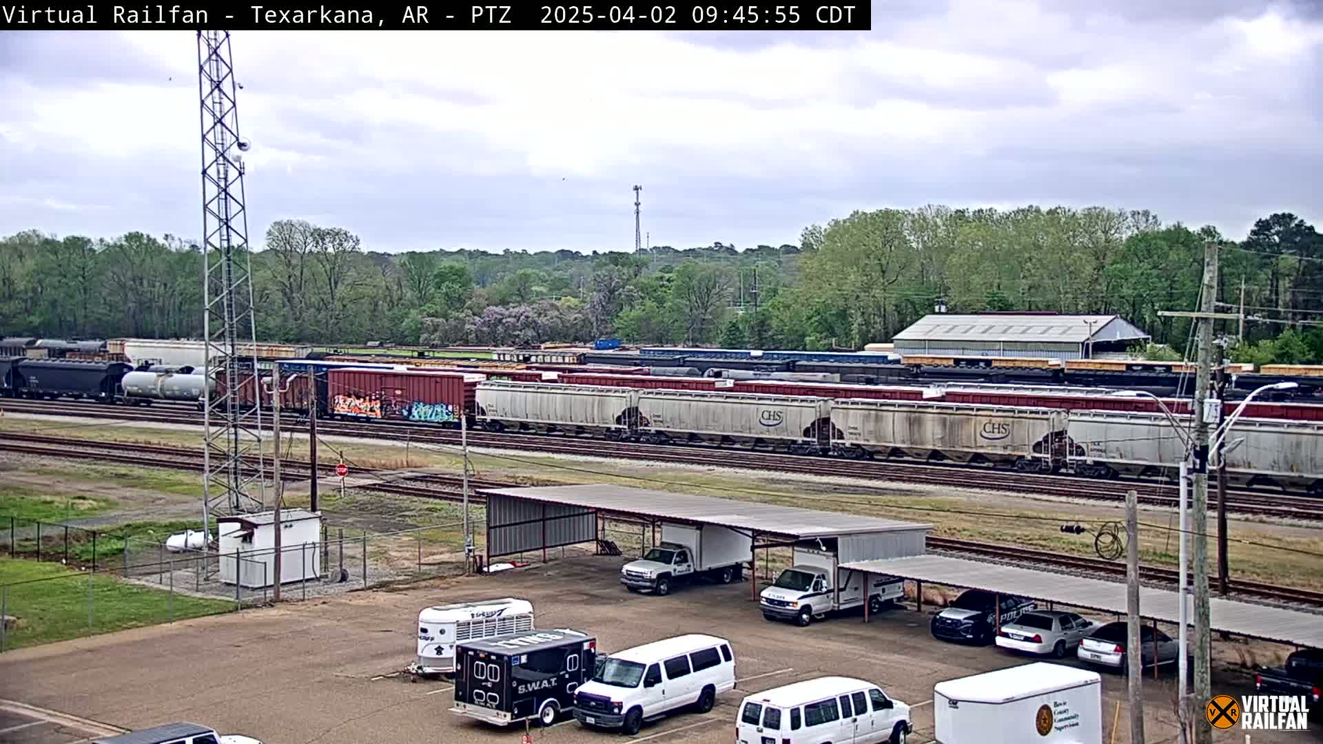 A long freight train sits on railroad tracks next to a parking lot with several vehicles under a covered structure, all under a partly cloudy sky.