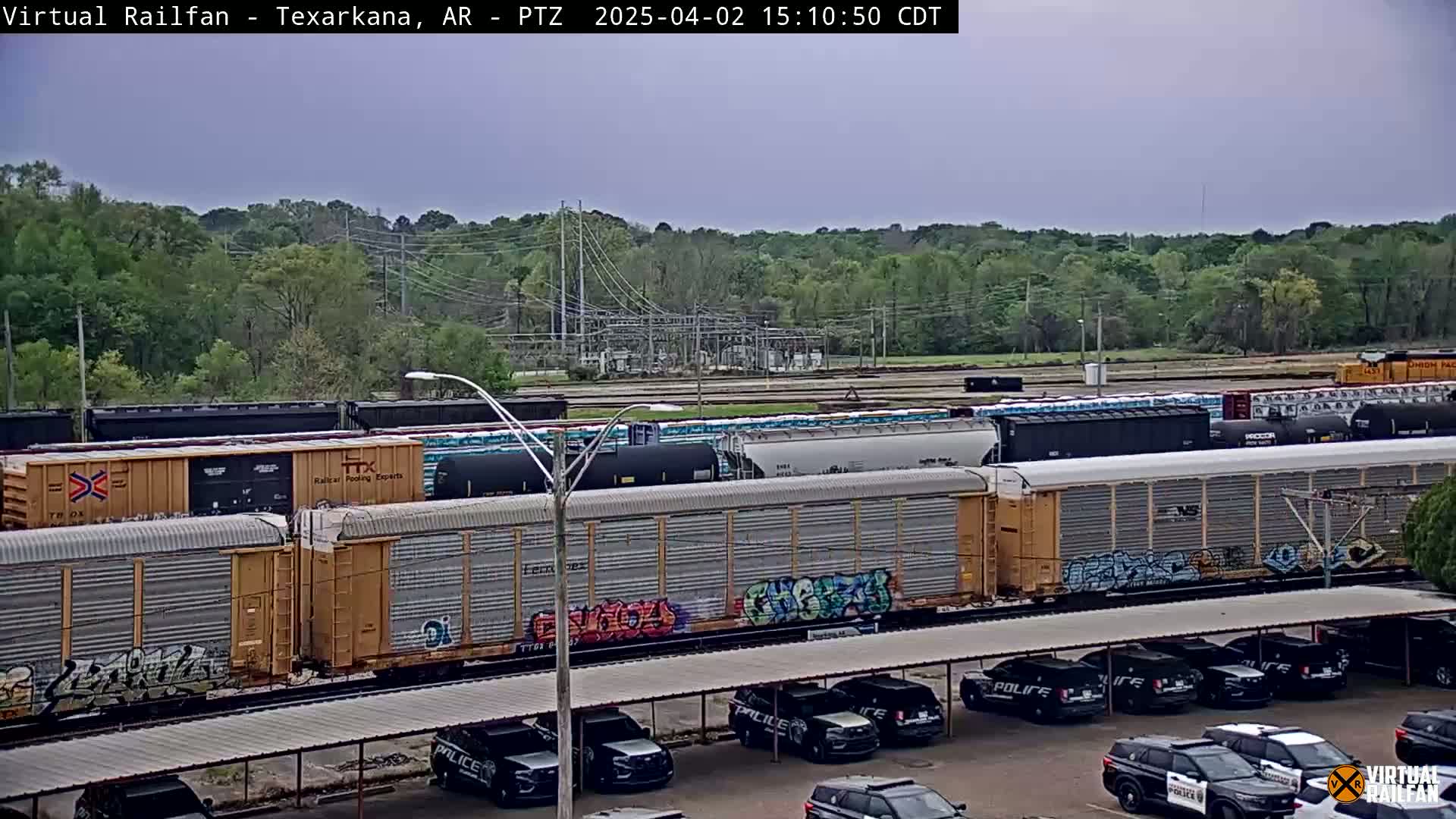 A freight train sits near a parking lot full of police vehicles under an overcast sky, with a treeline and electrical substation visible in the background.