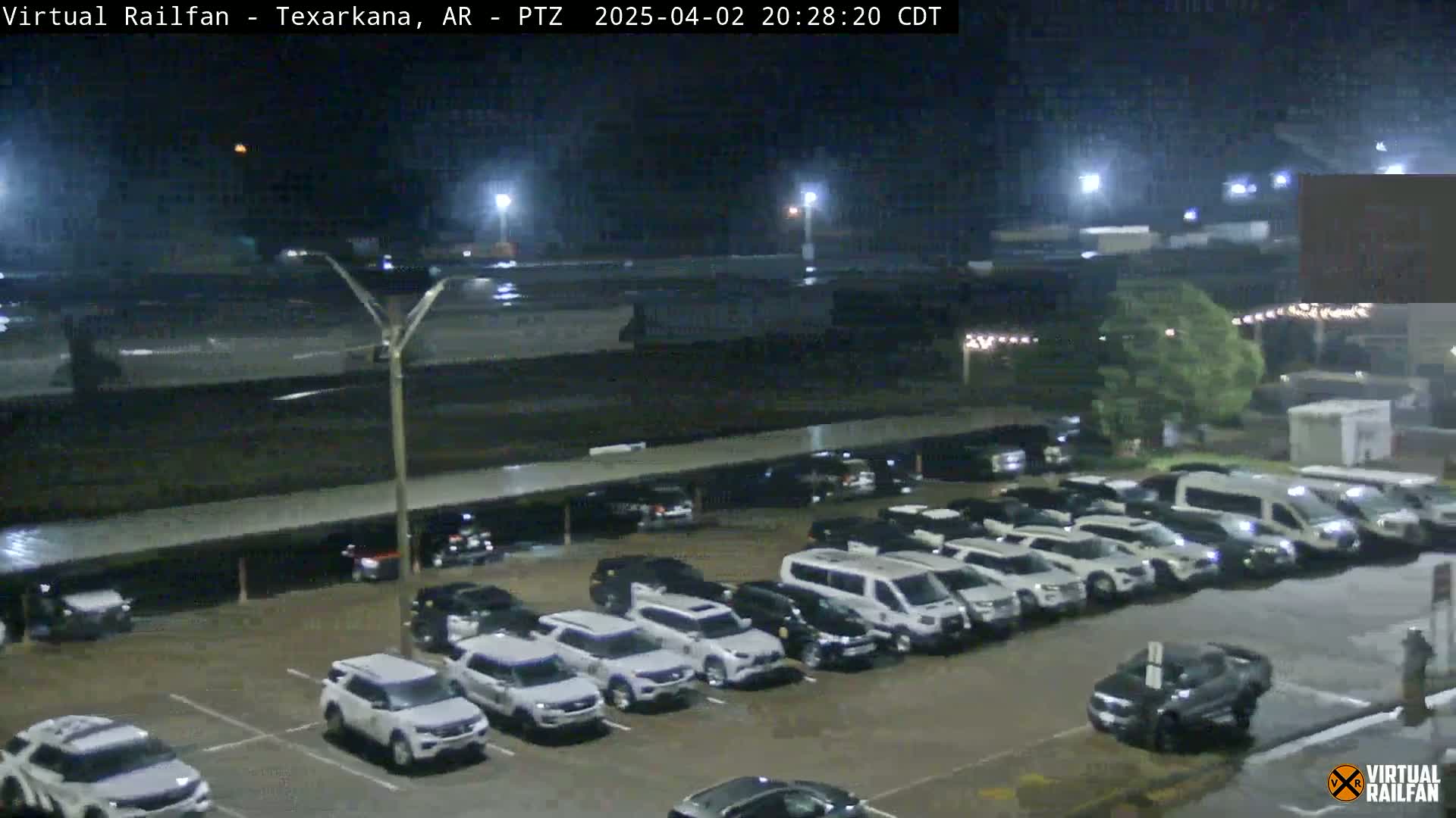 A nighttime, rain-slicked parking lot filled with mostly white SUVs and vans under dim lighting.