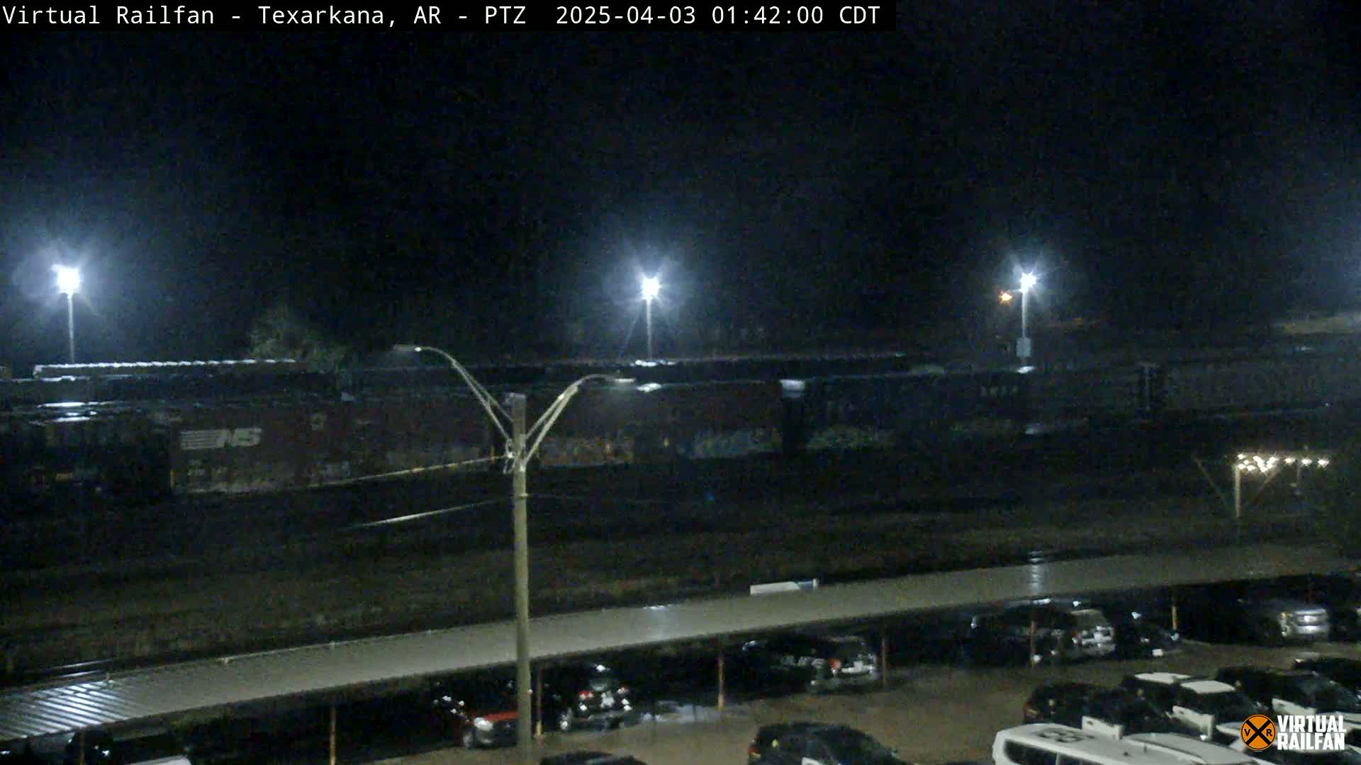 At night, under bright lights, a long train of boxcars sits parked next to a parking lot filled with cars.