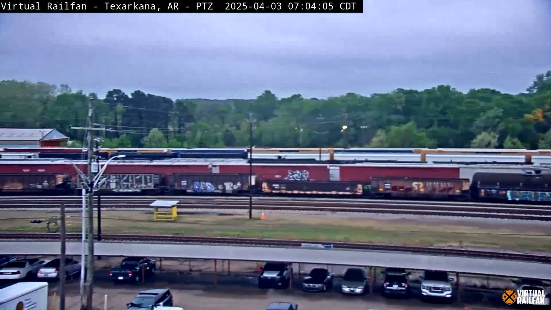 A long freight train sits on railroad tracks next to a parking lot under an overcast sky.