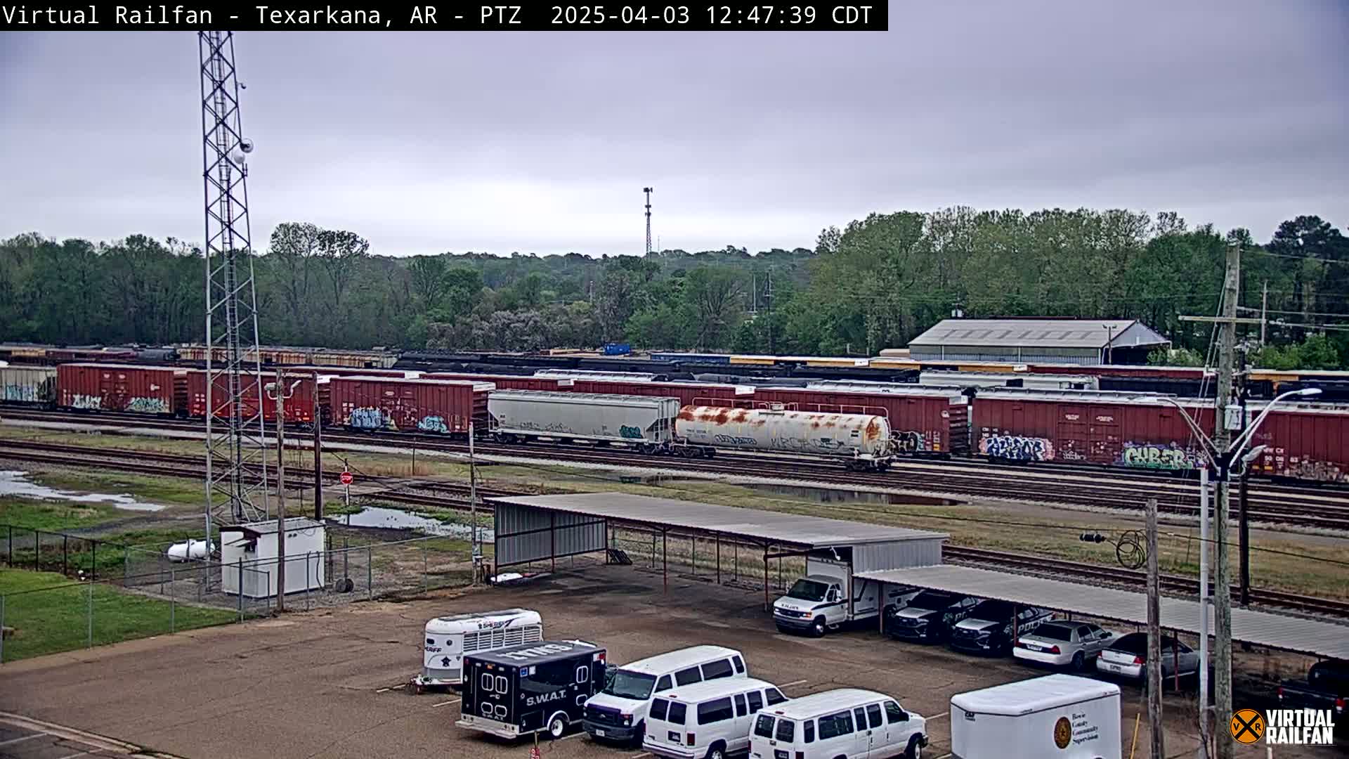 A long freight train sits on railroad tracks under an overcast sky, with several vehicles parked under a covered structure nearby.