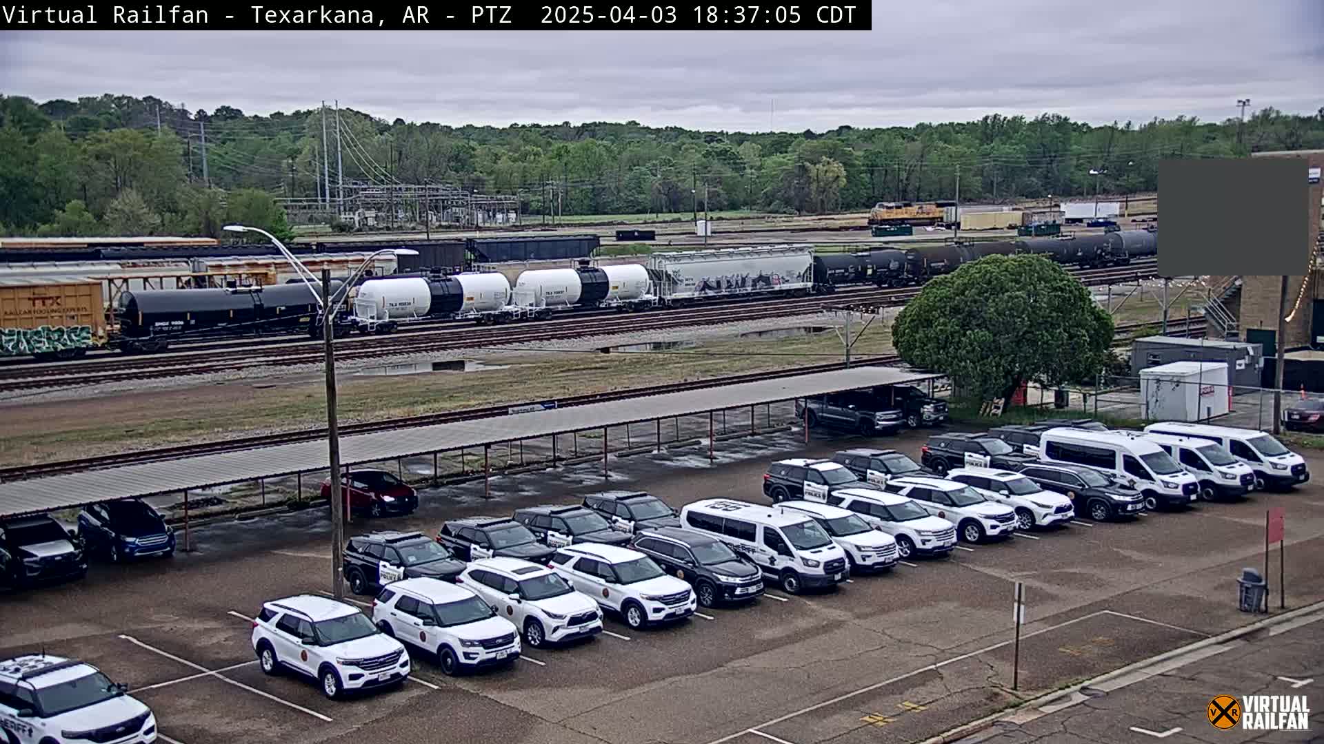 A large parking lot filled with mostly white SUVs is situated near a railway with several freight cars and a distant locomotive under an overcast sky.