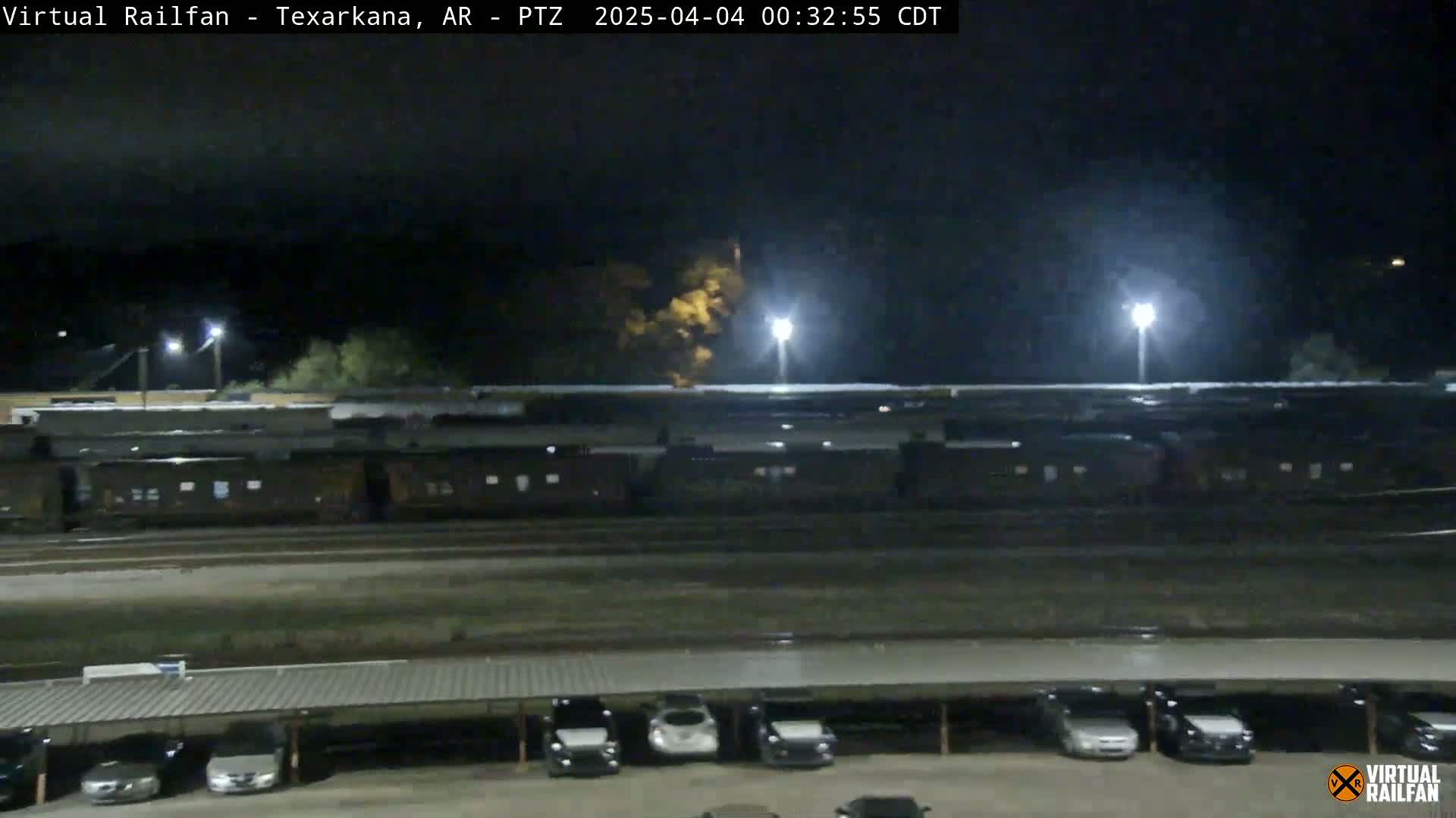A nighttime view shows a long freight train parked on railway tracks next to a parking lot with cars under a covered structure.
