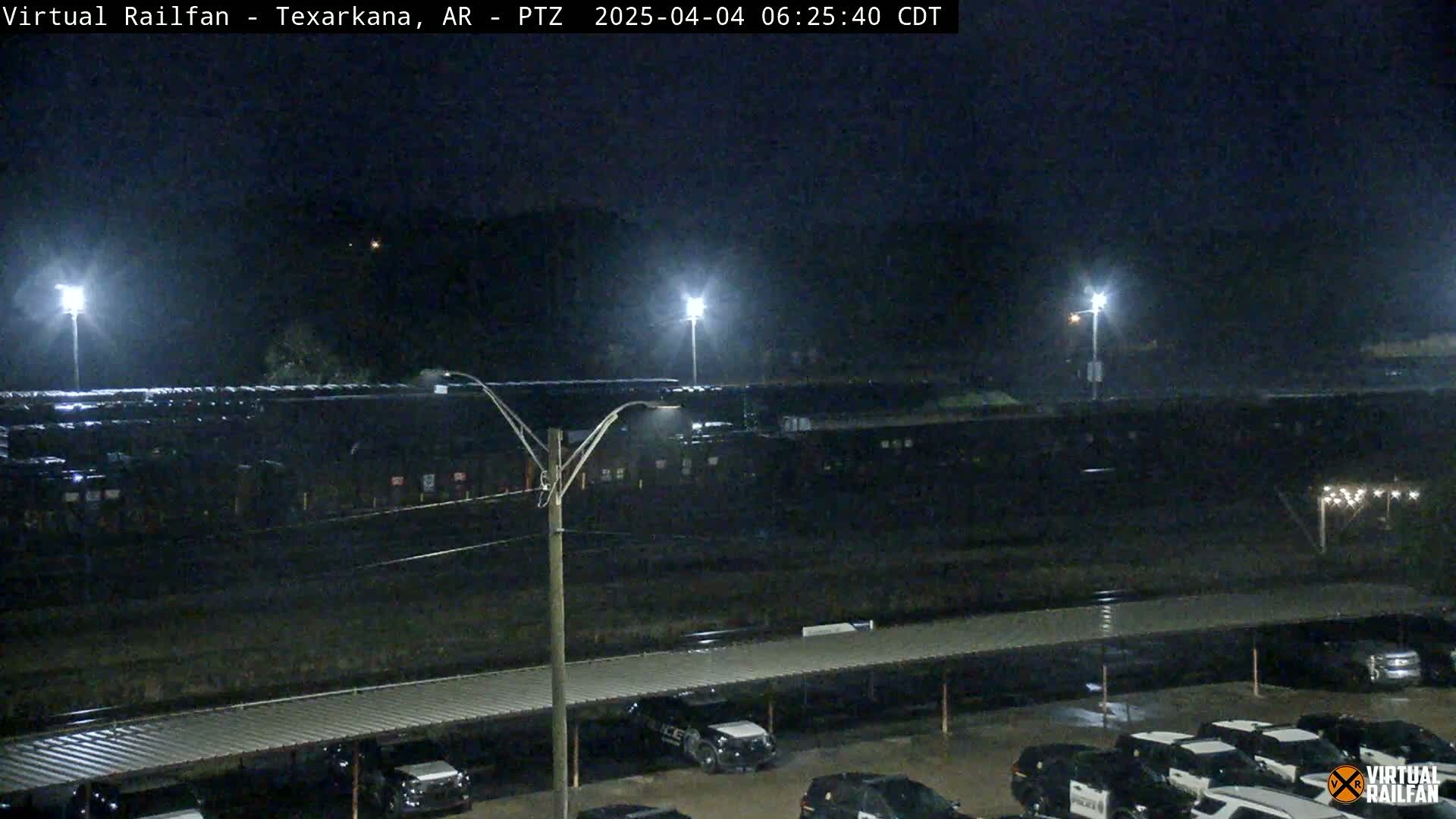 At night, under a dark sky, a long train sits in a rail yard next to a parking lot with several police cars parked under a covered structure.