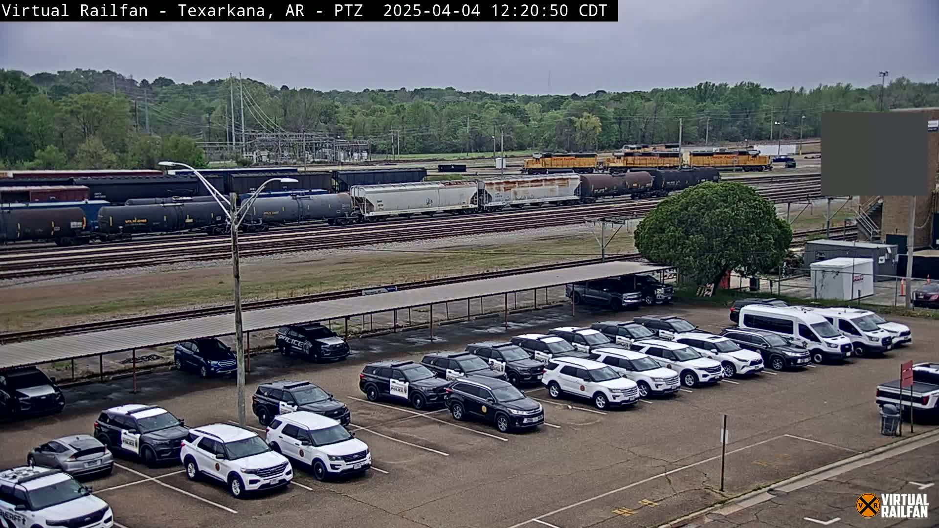 A train of freight cars sits on a rail line next to a parking lot full of police SUVs under an overcast sky.