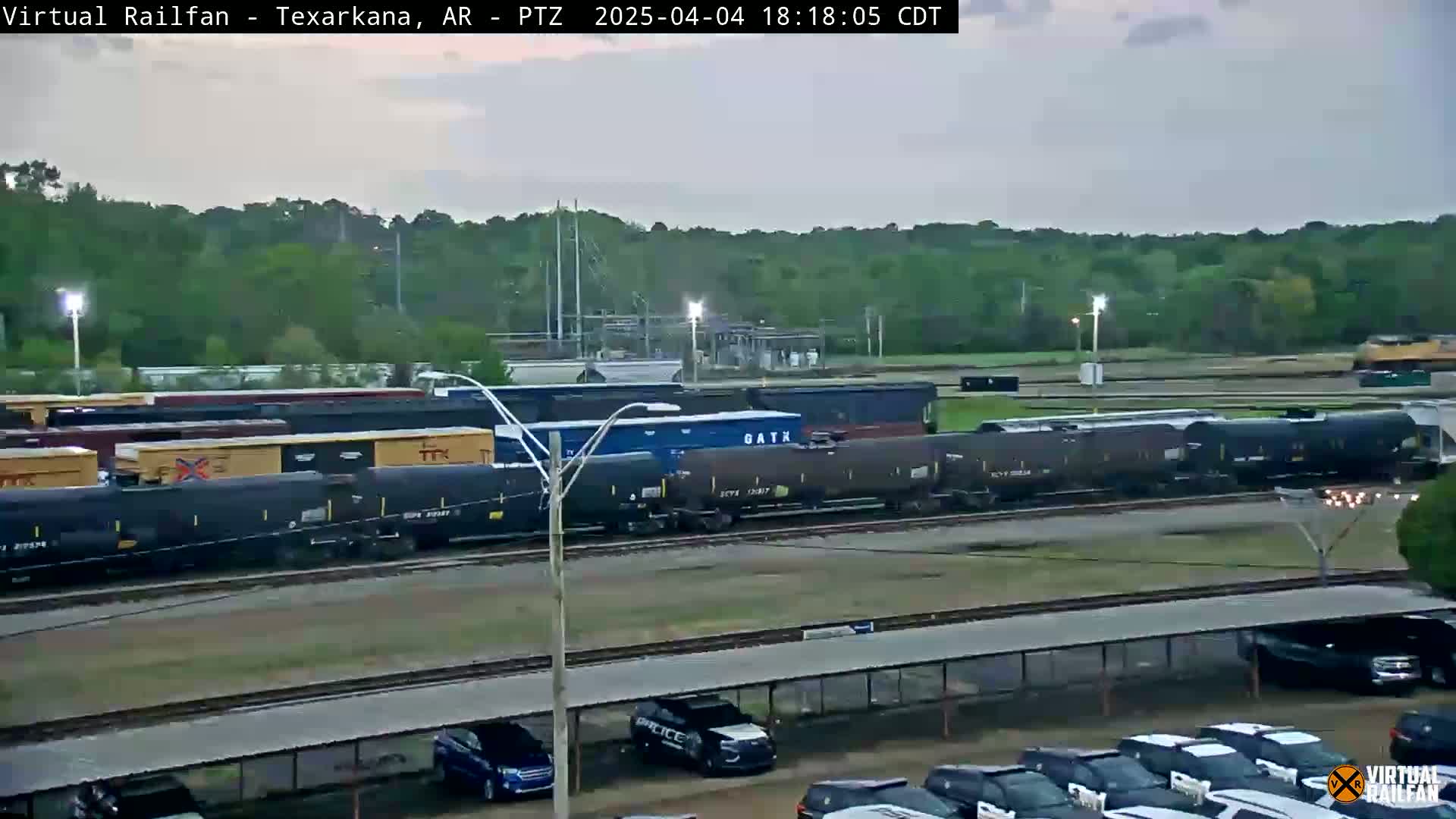 A long train of dark-colored tanker cars sits on a railway track in front of a line of parked cars under an overcast sky.