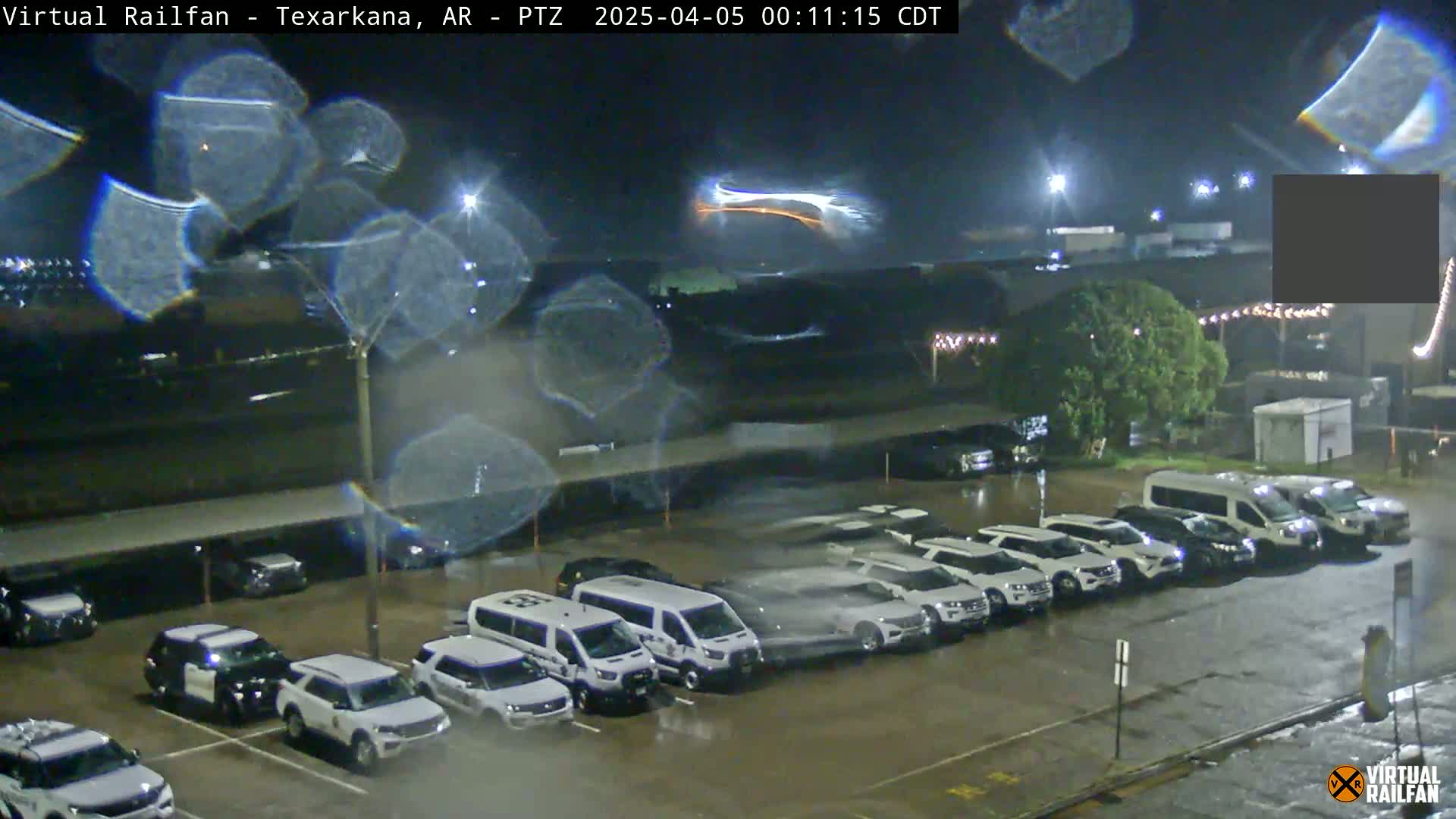 A nighttime, rain-slicked parking lot filled with mostly white SUVs and vans, with a distant, bright streak of light in the sky.