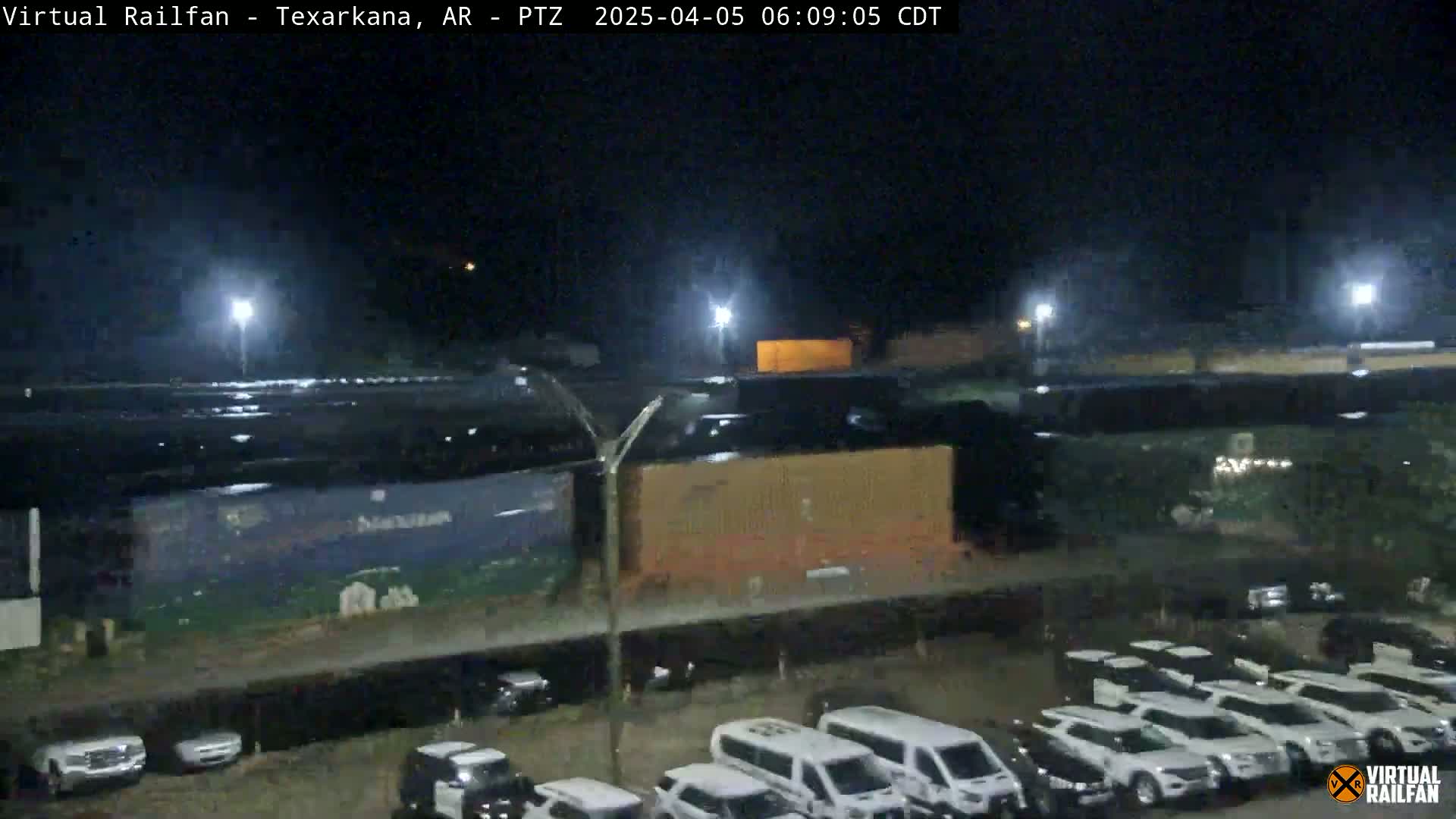 At night, under bright artificial lights, a train car and numerous white vehicles are parked in a large, mostly empty lot.
