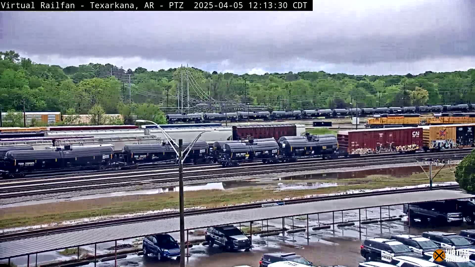 A large number of railway cars are parked on several tracks under an overcast sky, with puddles in a parking lot in the foreground.