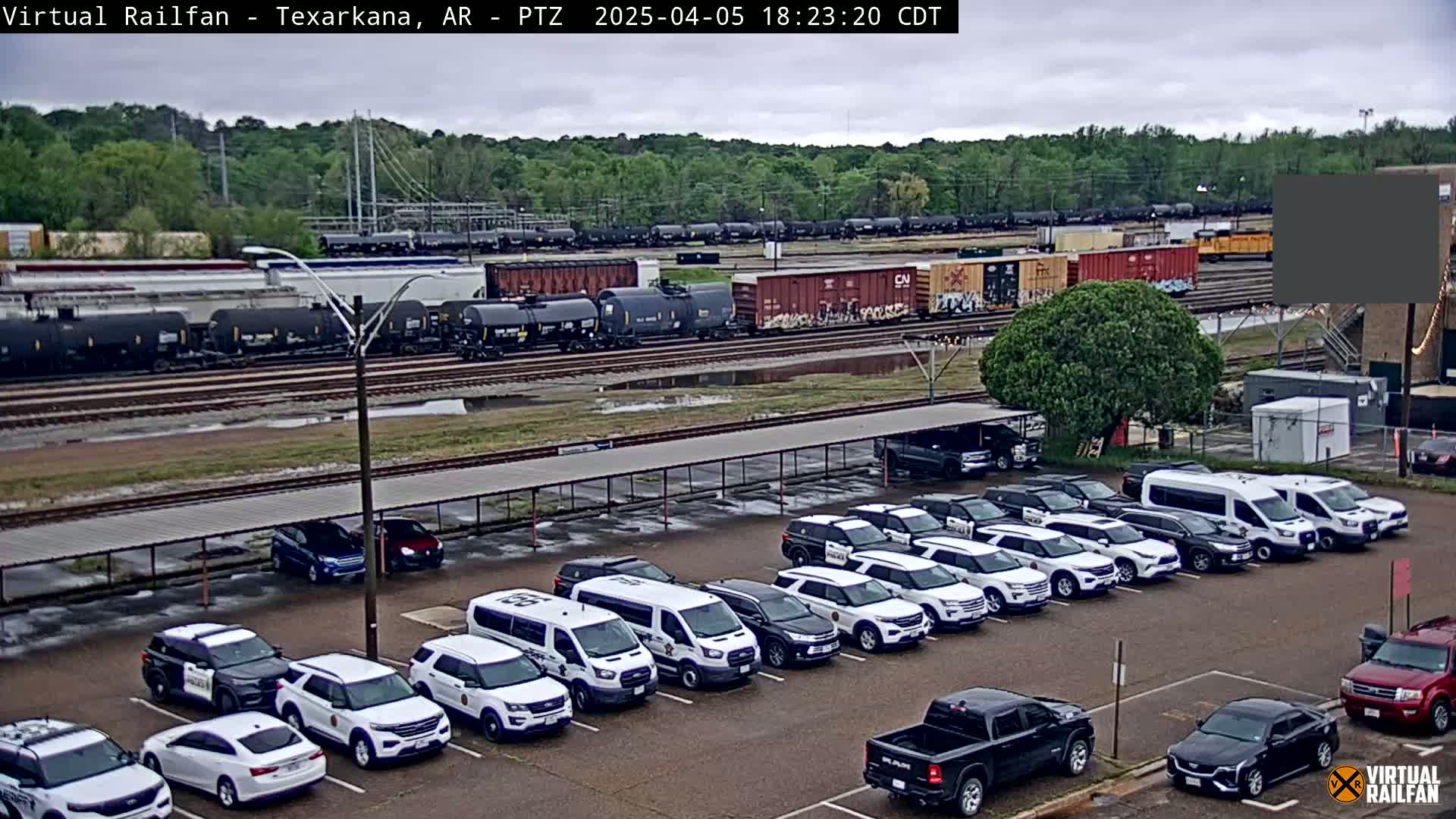 A large parking lot containing numerous white police vehicles sits under an overcast sky next to a railroad yard filled with freight cars.