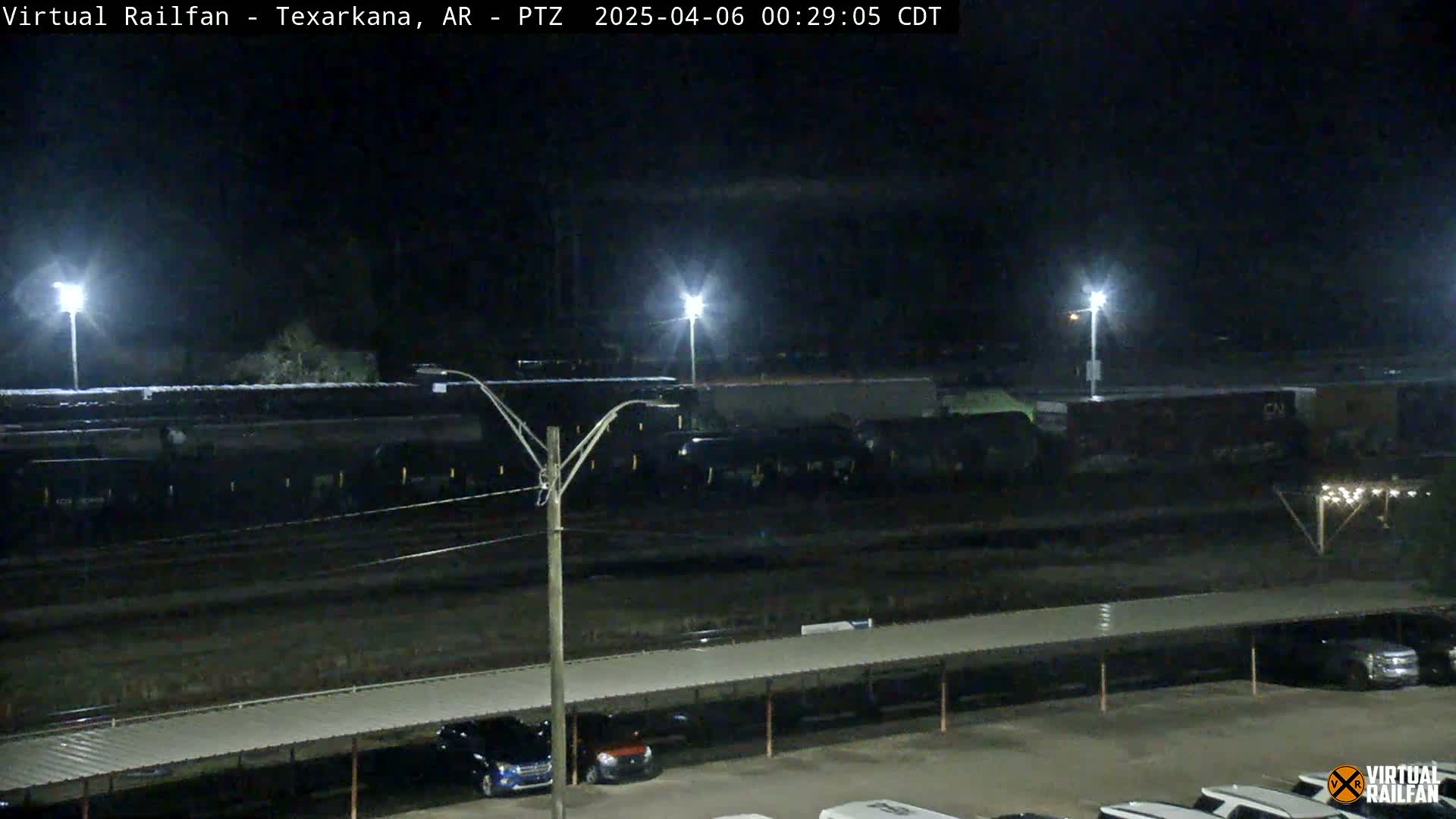 A long freight train sits on railroad tracks at night under bright lights, next to a parking lot with cars parked under a covered structure.