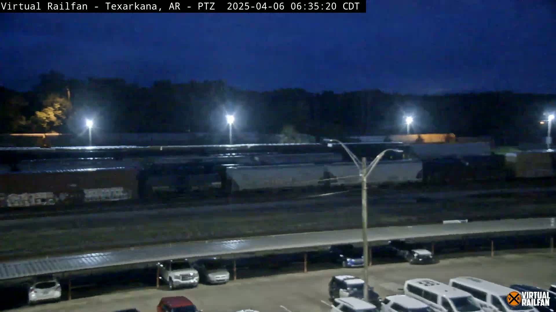 Under a dark night sky, a train sits in a rail yard next to a parking lot with several cars and vans parked under a covered structure.