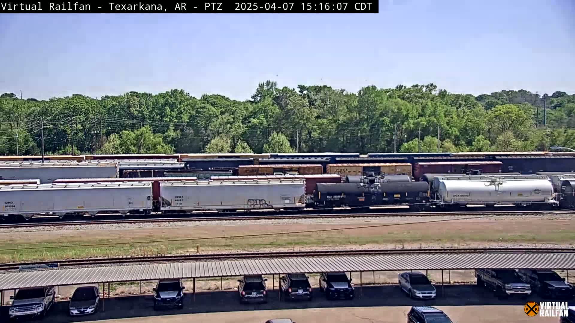 A long freight train sits on the tracks under a clear, sunny sky, parked near a lot with several cars under a covered structure.