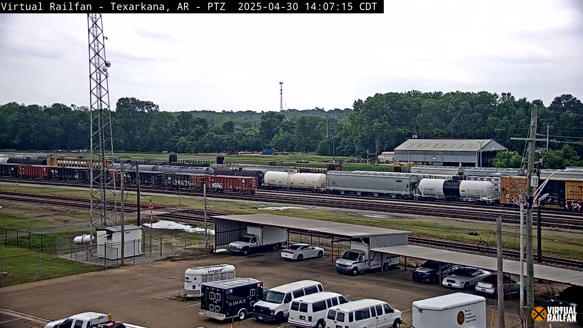 A long freight train sits on railroad tracks under an overcast sky, with several vehicles parked under a covered structure nearby.