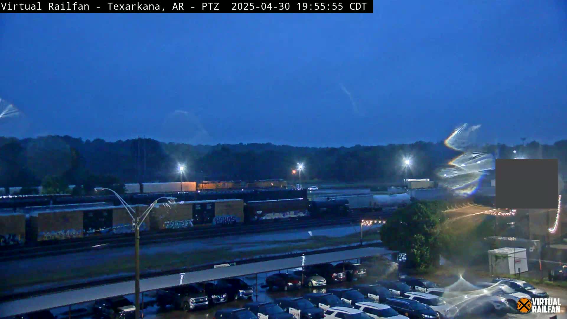 A long freight train sits under a dark blue, rainy twilight sky next to a parking lot.