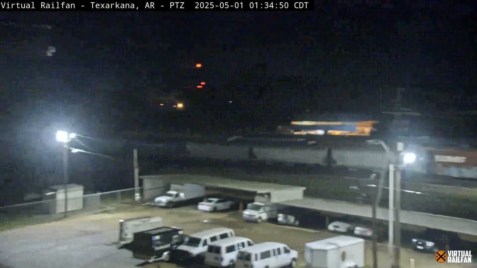 A nighttime view of a parking lot with several vans and trucks, under bright lights, near a railway line.
