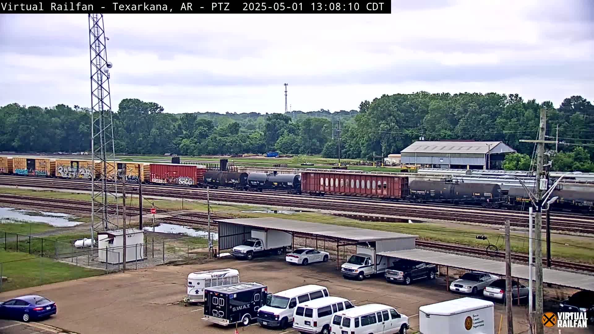 A long freight train passes by a parking lot with several vehicles under a covered structure, all situated near a line of trees under an overcast sky.
