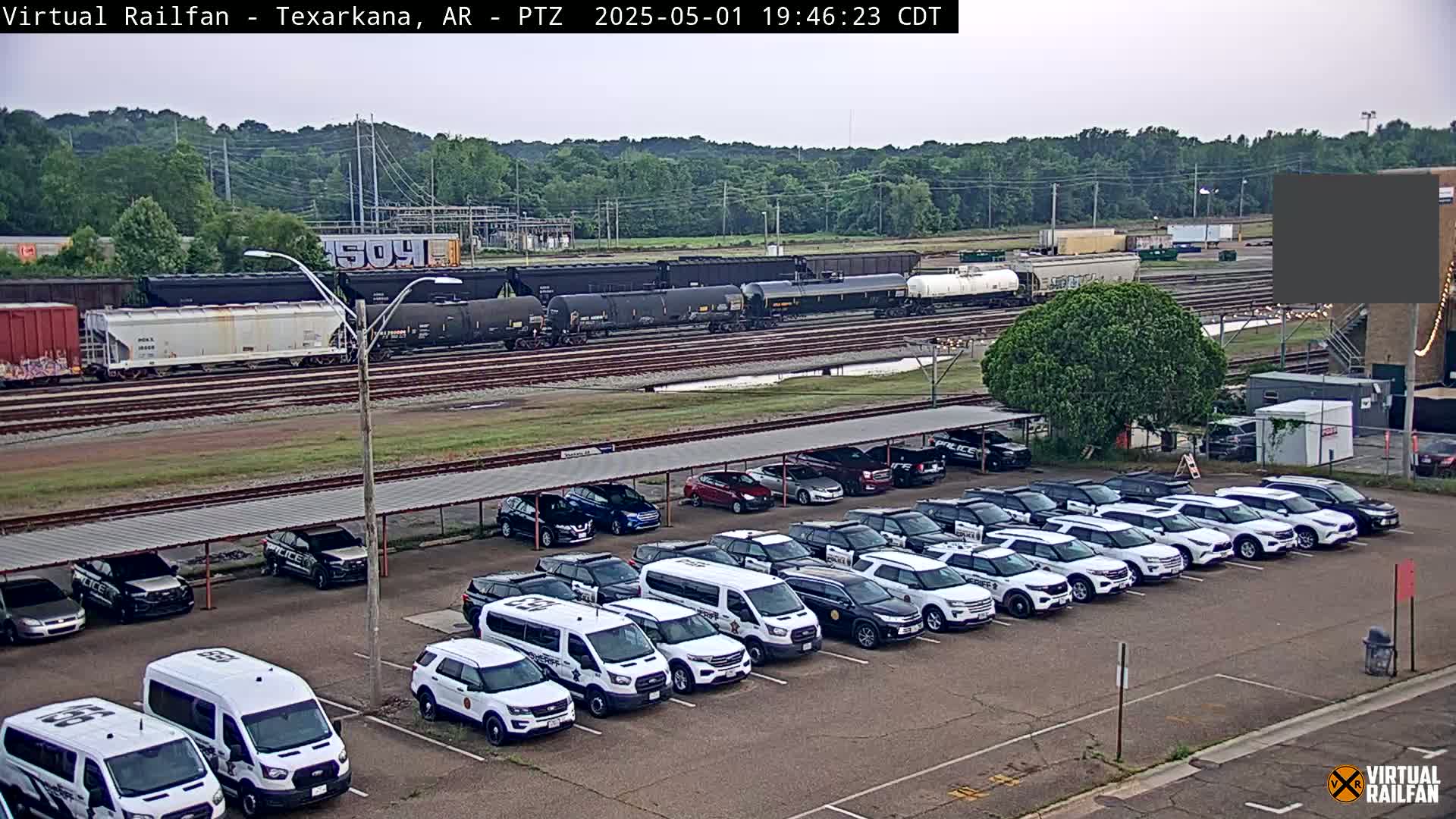 A train of freight cars sits on railway tracks next to a parking lot full of mostly white police vehicles under a covered structure, all under an overcast sky.