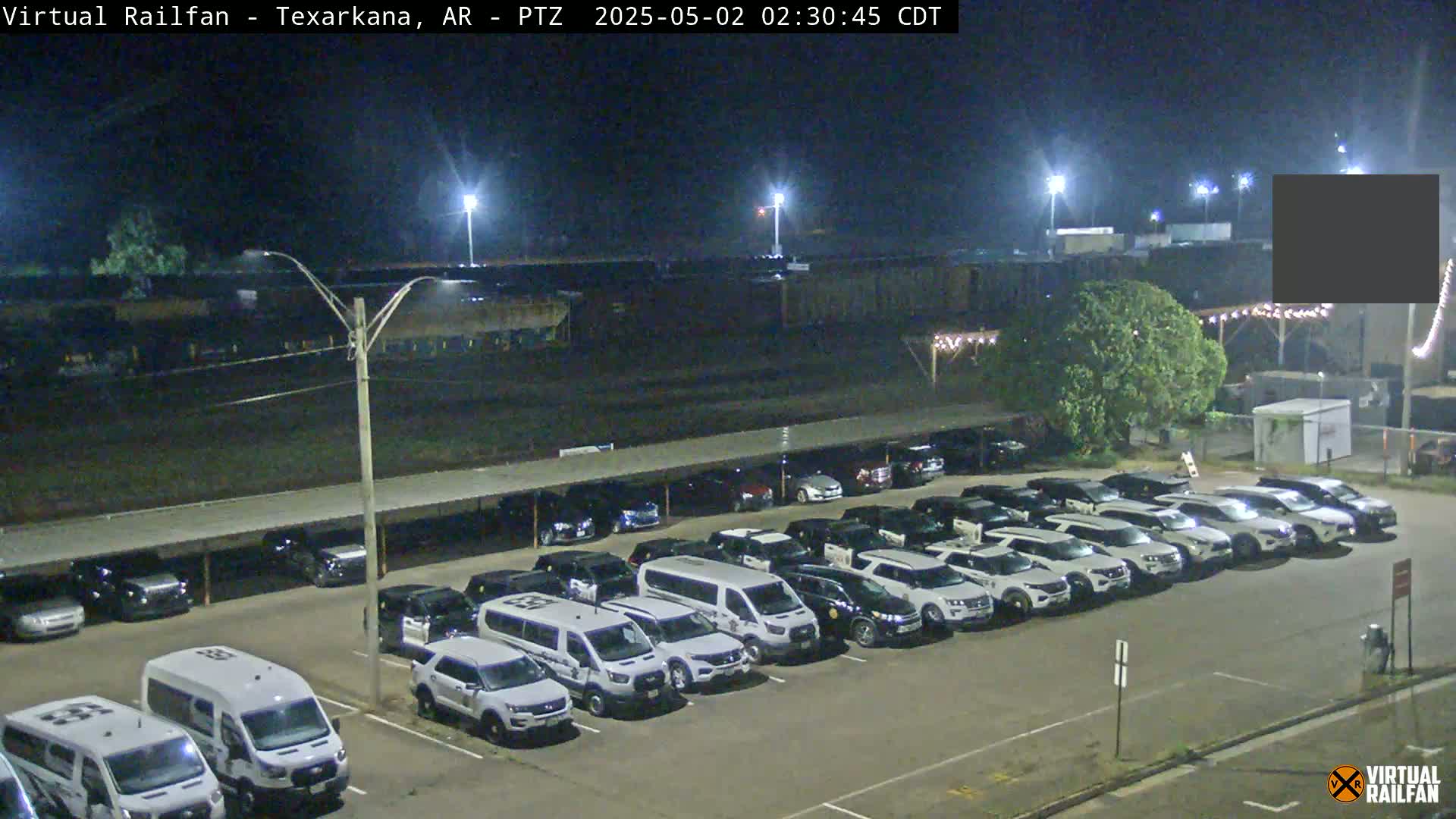A nighttime view of a parking lot filled with mostly white SUVs and vans under a covered structure, next to a large dark building and a tree.