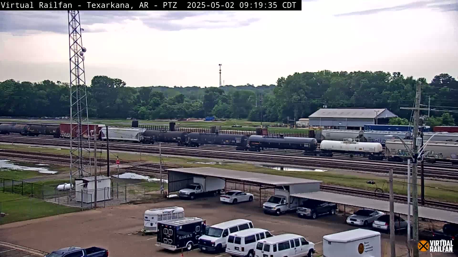 A long freight train sits on the tracks under an overcast sky, with several vehicles parked under a covered structure in the foreground.