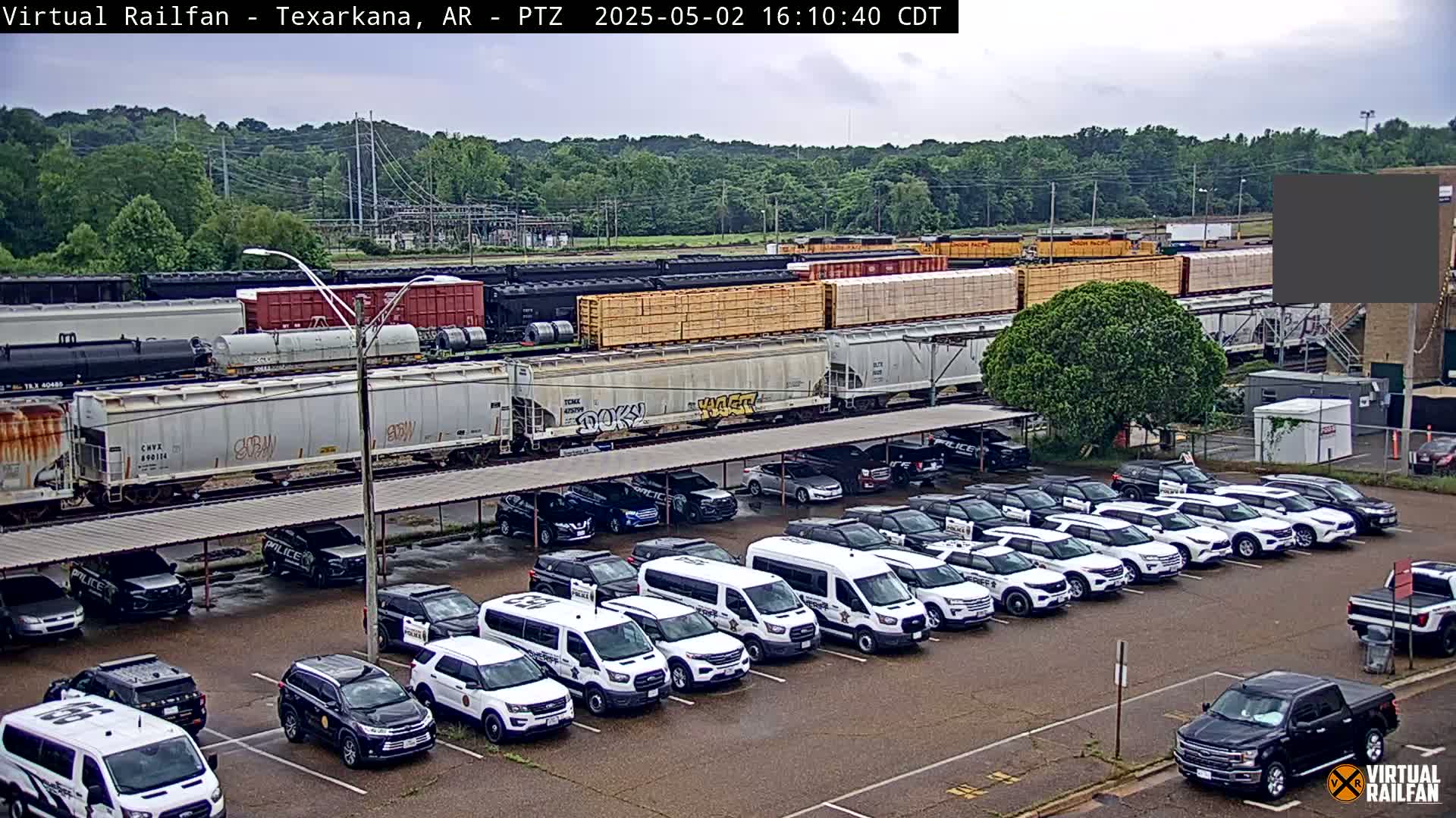 Under an overcast sky, a long freight train is parked beside a lot full of mostly white police vehicles under a covered structure.