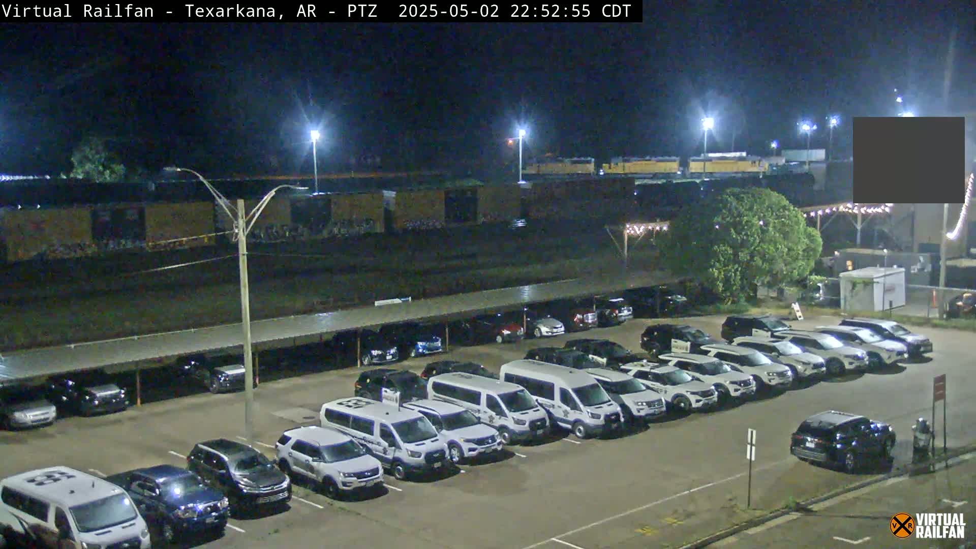 A nighttime view of a parking lot filled with cars and vans, situated near a railway with freight cars, under clear skies.