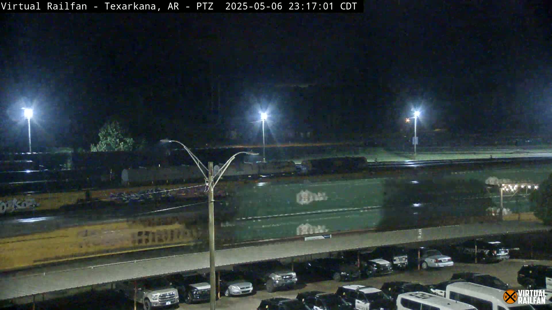 A long freight train passes by a parking lot at night under wet conditions.