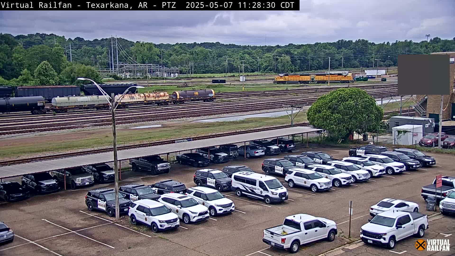 Under an overcast sky, a large parking lot filled with police vehicles sits next to a railway yard with several train cars and locomotives.