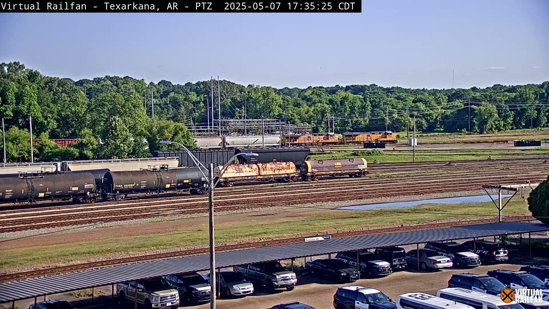 A sunny day reveals a railway yard with several freight cars and locomotives, along with a parking lot full of parked vehicles under a covered structure, all set against a backdrop of lush green trees.