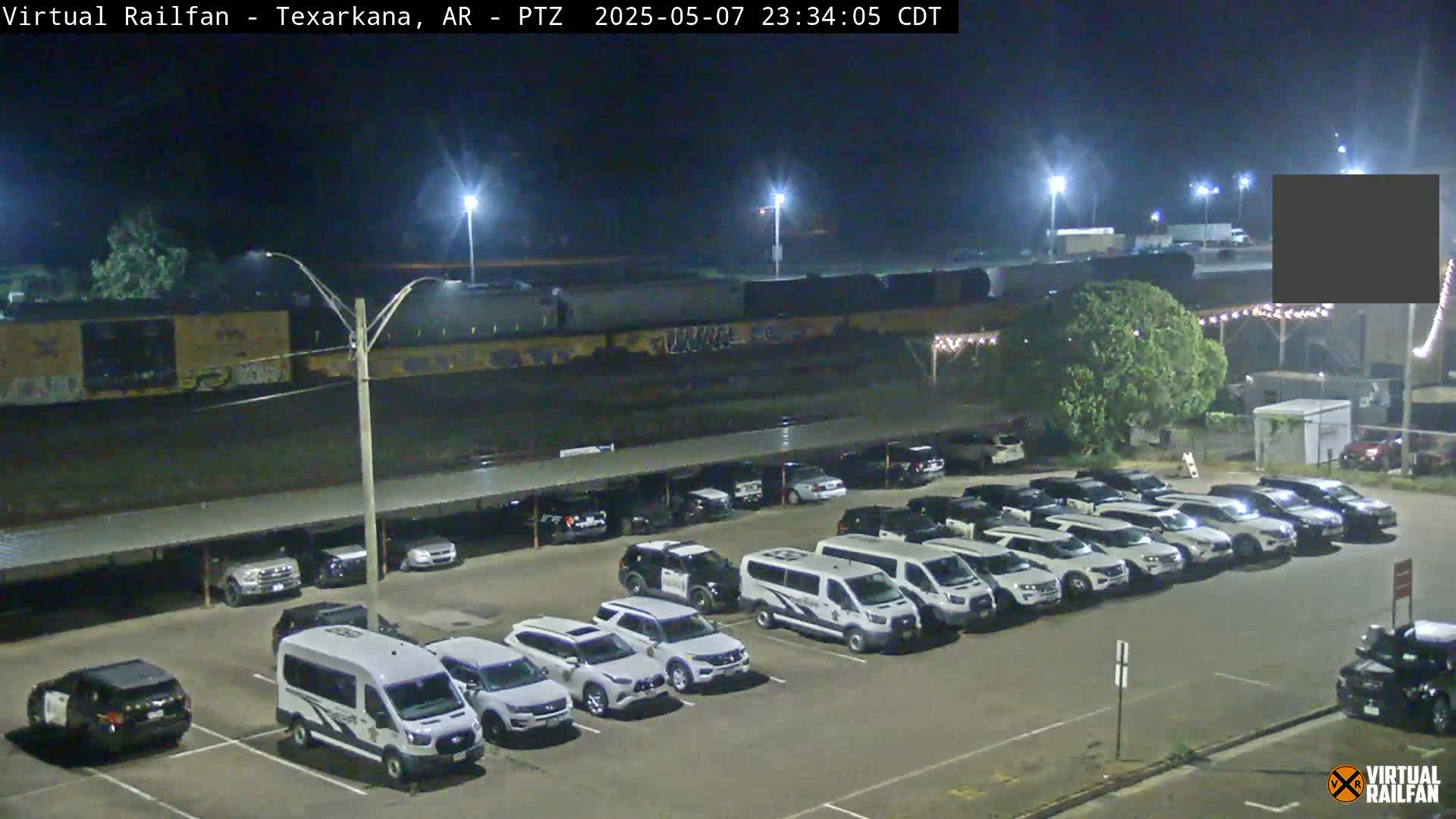 A long freight train sits behind a parking lot full of mostly white SUVs and vans at night under bright lights.