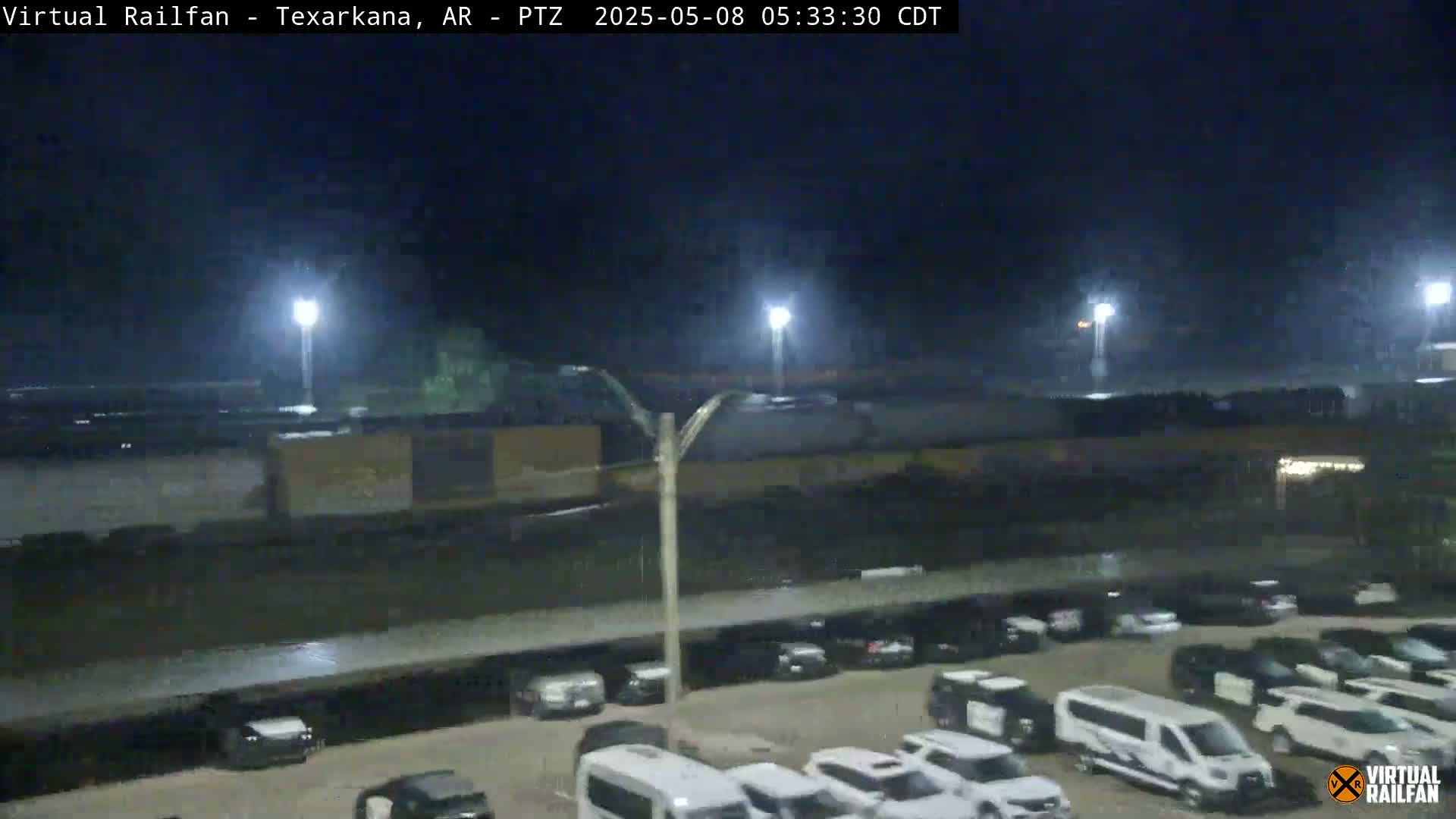 At night, under a dark sky, a long freight train passes over a parking lot full of vehicles, wet from recent rain.