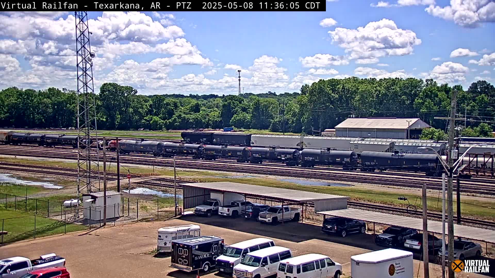 A long freight train sits on railroad tracks under a partly cloudy sky, with several vehicles parked under a covered structure nearby.