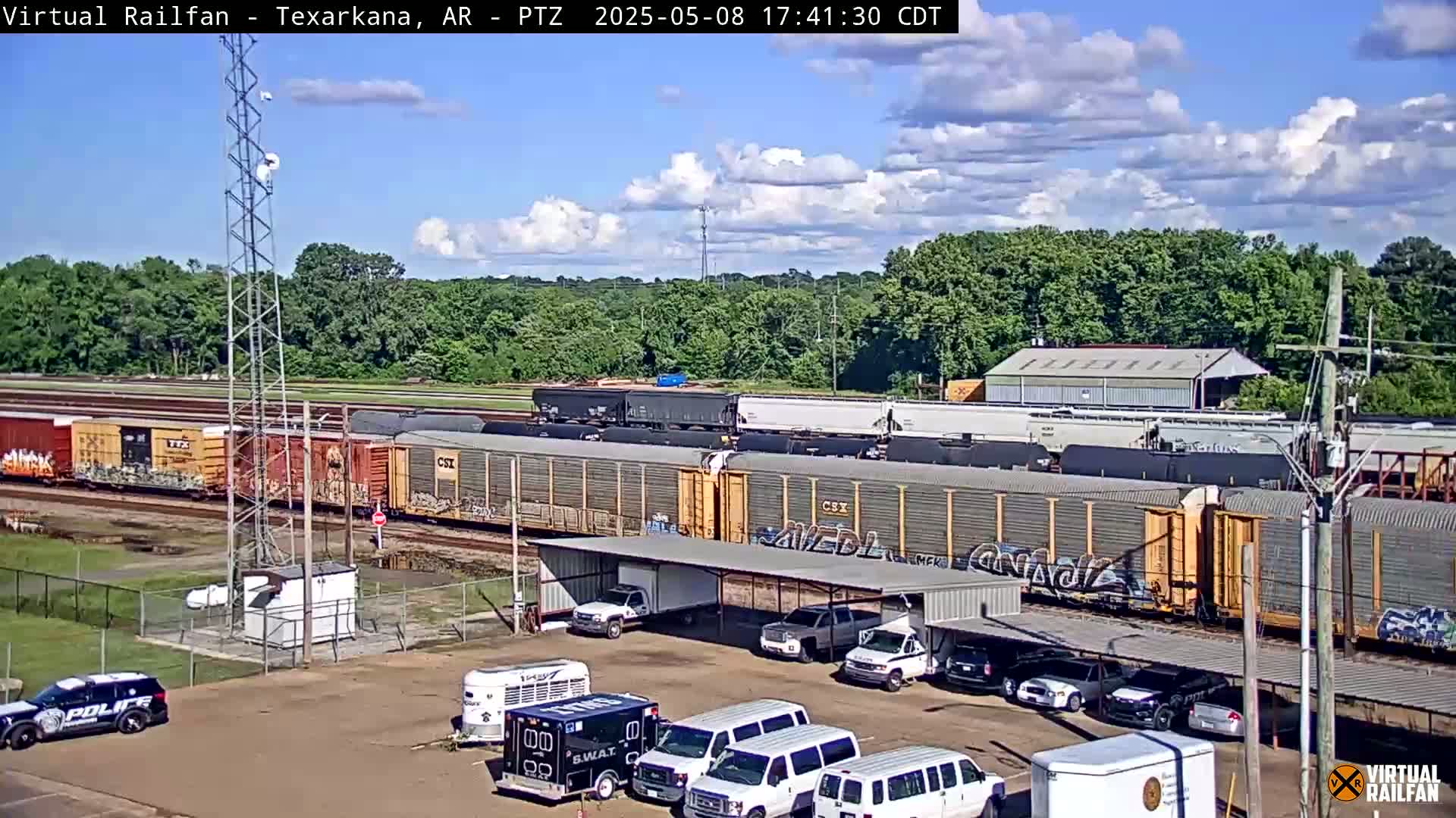 A long freight train sits on the tracks under a partly cloudy sky, parked next to a lot with several parked cars and vans, including police vehicles.