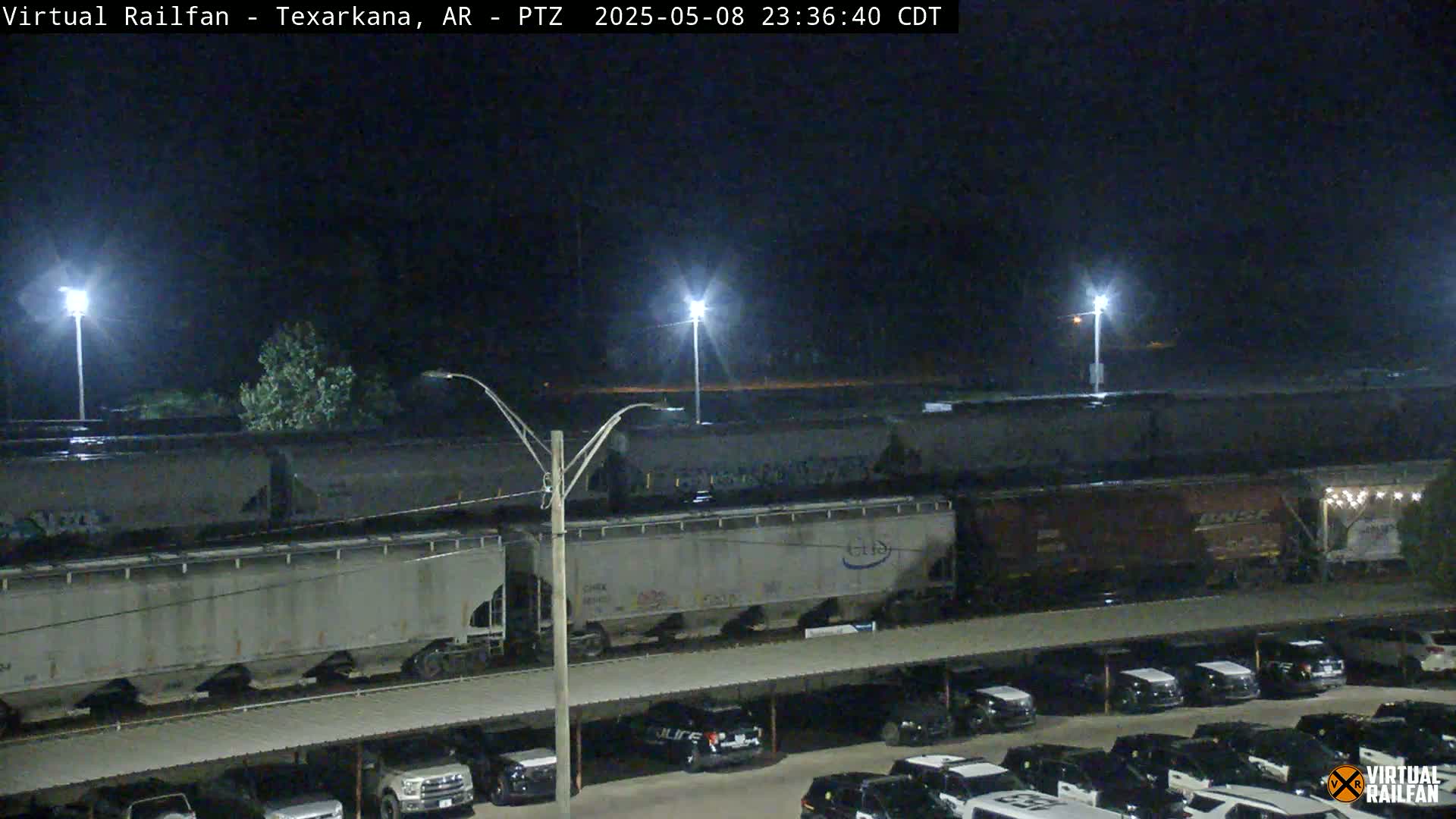 A long freight train sits under nighttime conditions near a parking lot filled with cars.