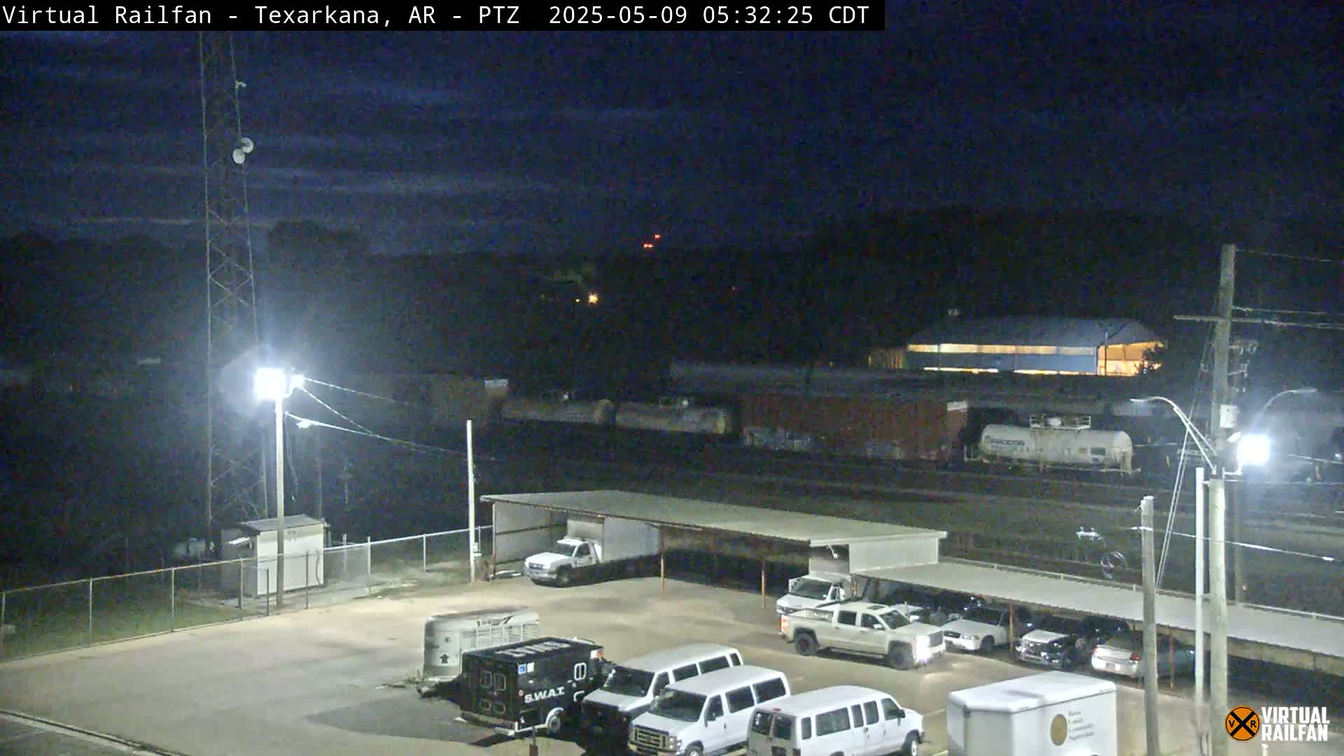 A nighttime view of a railway yard with several parked vehicles under a covered structure, a train in the background, and a tall tower in the foreground under a dark, mostly clear sky.
